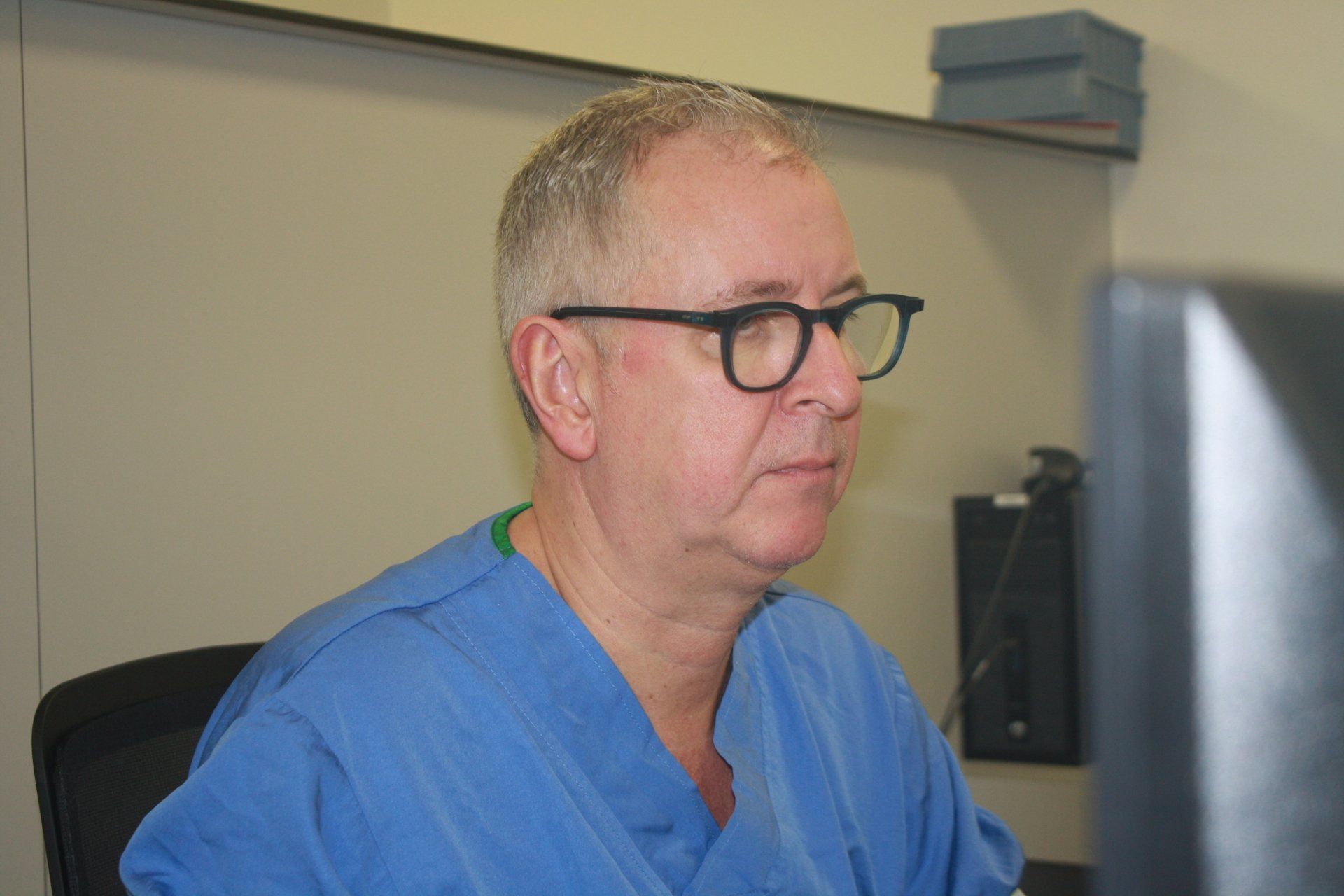 Een man in een blauwe operatiekleding en met een bril zit aan een bureau en kijkt naar een computermonitor in een kantoor.
