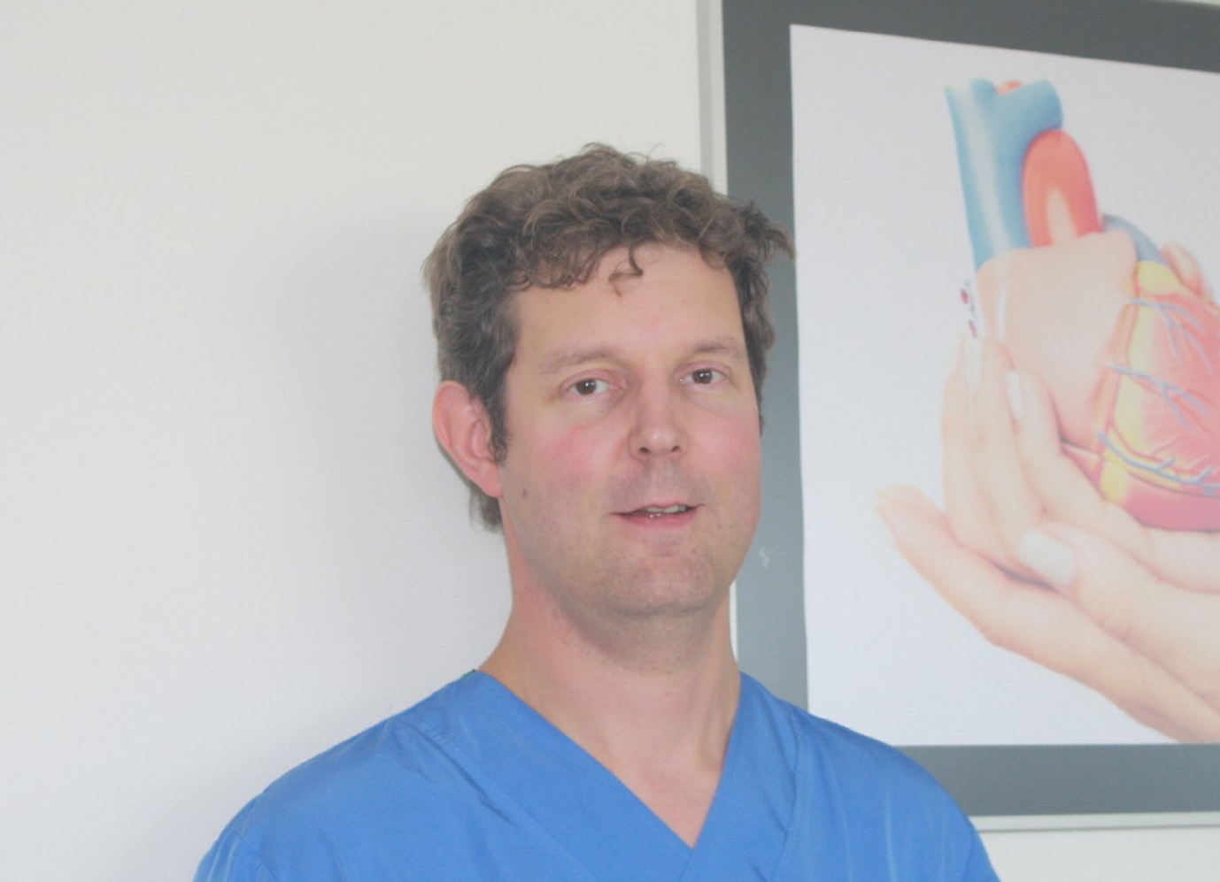 Man in blauwe operatiekleding staat binnen naast een poster met medische anatomie.