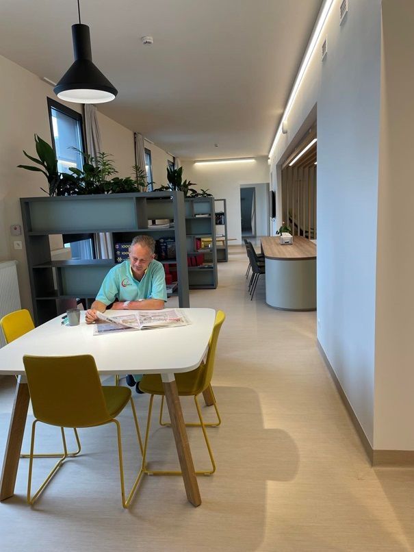 Een persoon zit aan een witte tafel in een lichte, moderne lounge en leest tijdschriften in de buurt van boekenplanken en potplanten.