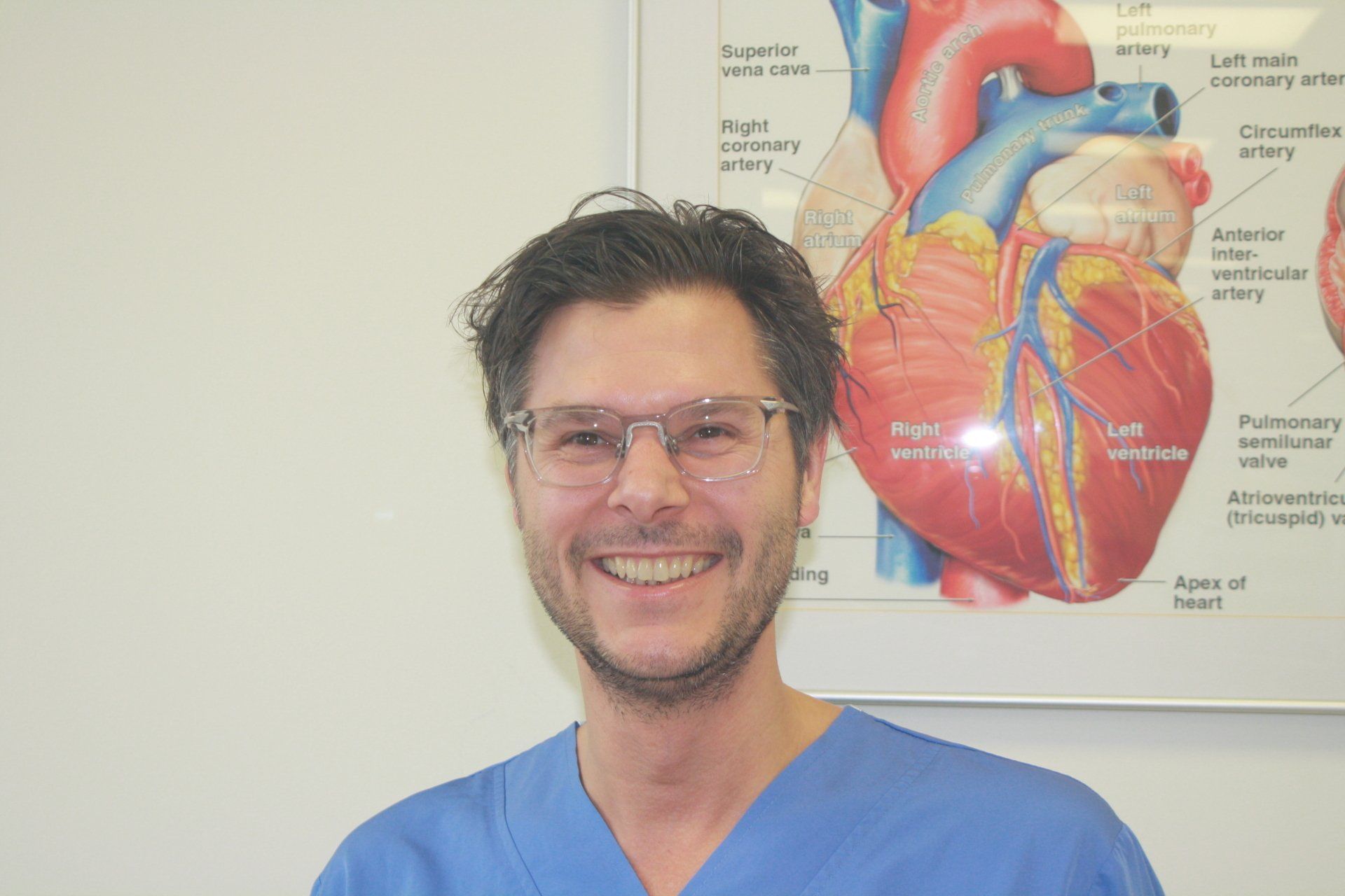 Een glimlachend persoon in een blauwe operatiekleding staat voor een poster met een afbeelding van de anatomie van het hart.