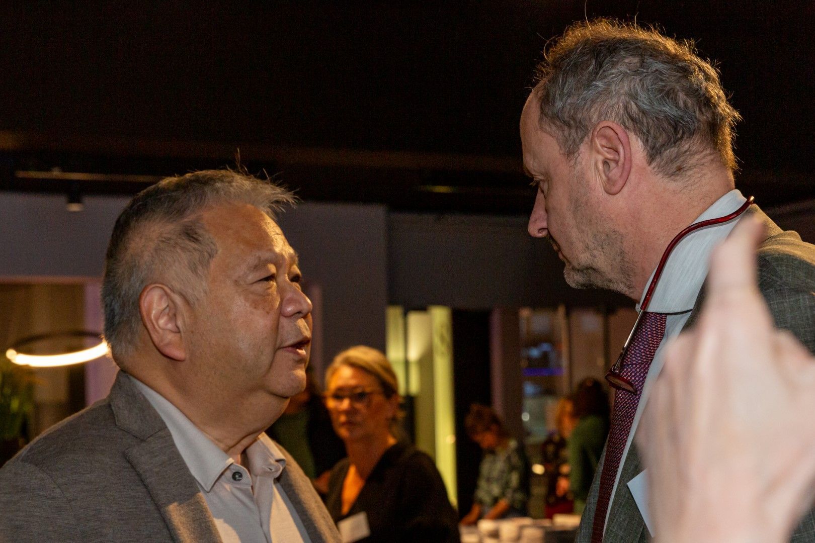 Twee mannen zitten tegenover elkaar in gesprek tijdens een evenement binnenshuis, met mensen op de achtergrond die wazig zijn weergegeven.