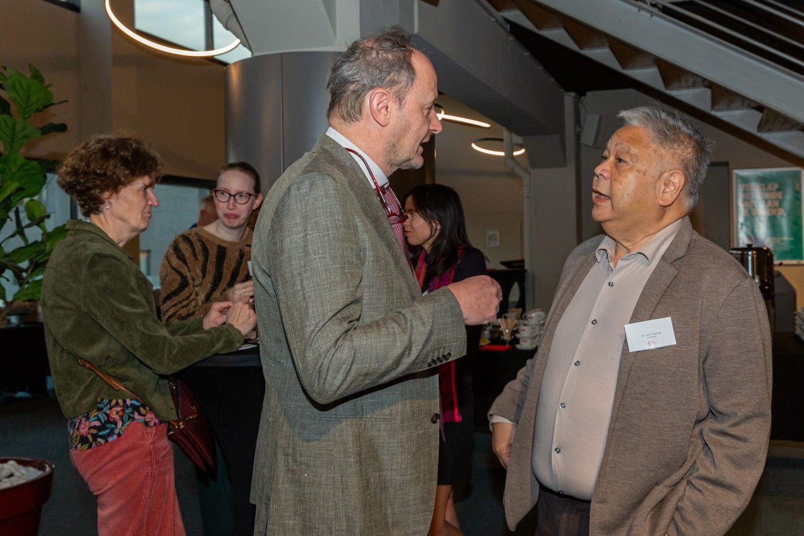 Twee mensen in pak praten met elkaar tijdens een sociale bijeenkomst binnenshuis, terwijl anderen op de achtergrond een gesprek voeren.