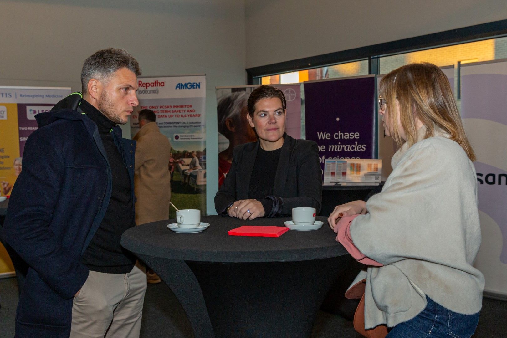 Drie mensen praten aan een ronde tafel in een stand op een evenement binnen, met spandoeken en een koffiekopje.
