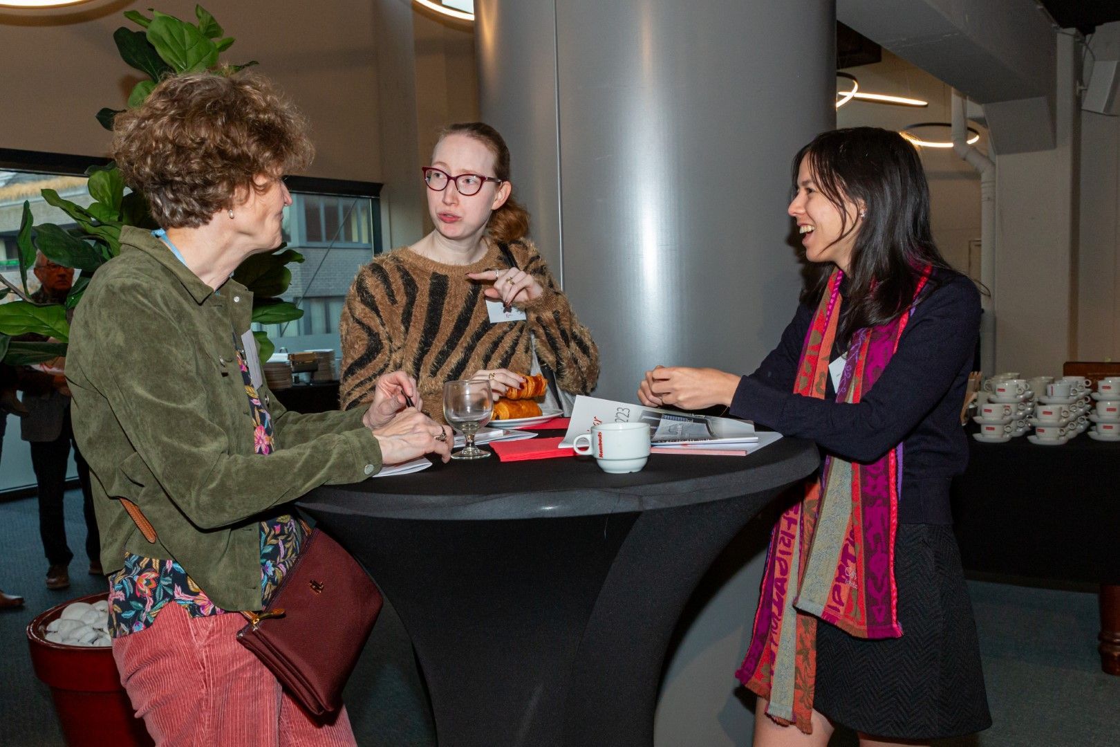 Drie mensen zitten te praten aan een hoge zwarte tafel, met hapjes en drankjes in een lichte, overdekte lounge.