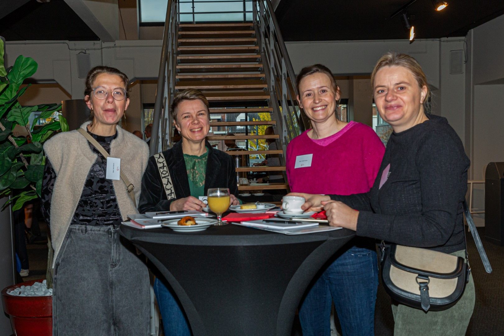 Vier mensen zitten aan een ronde tafel in een lobby, glimlachend met drankjes en borden onder een trap.