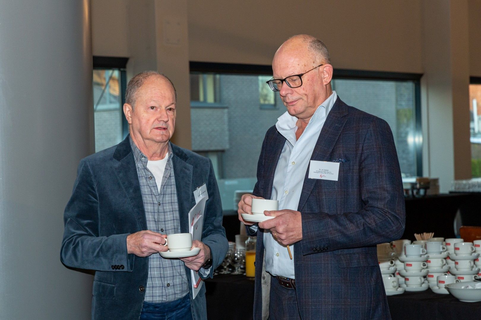 Twee mannen in colberts houden koffiekopjes vast aan een receptietafel, met op de achtergrond een stapel kopjes.