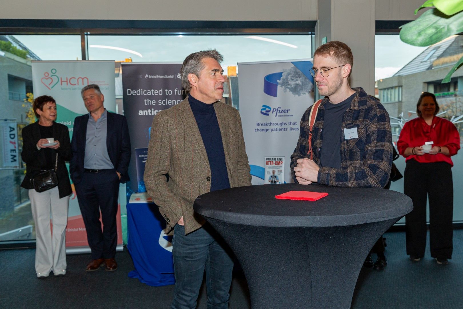 Twee mannen staan te praten naast een zwarte cocktailtafel in een conferentiestand, met spandoeken en deelnemers achter hen.
