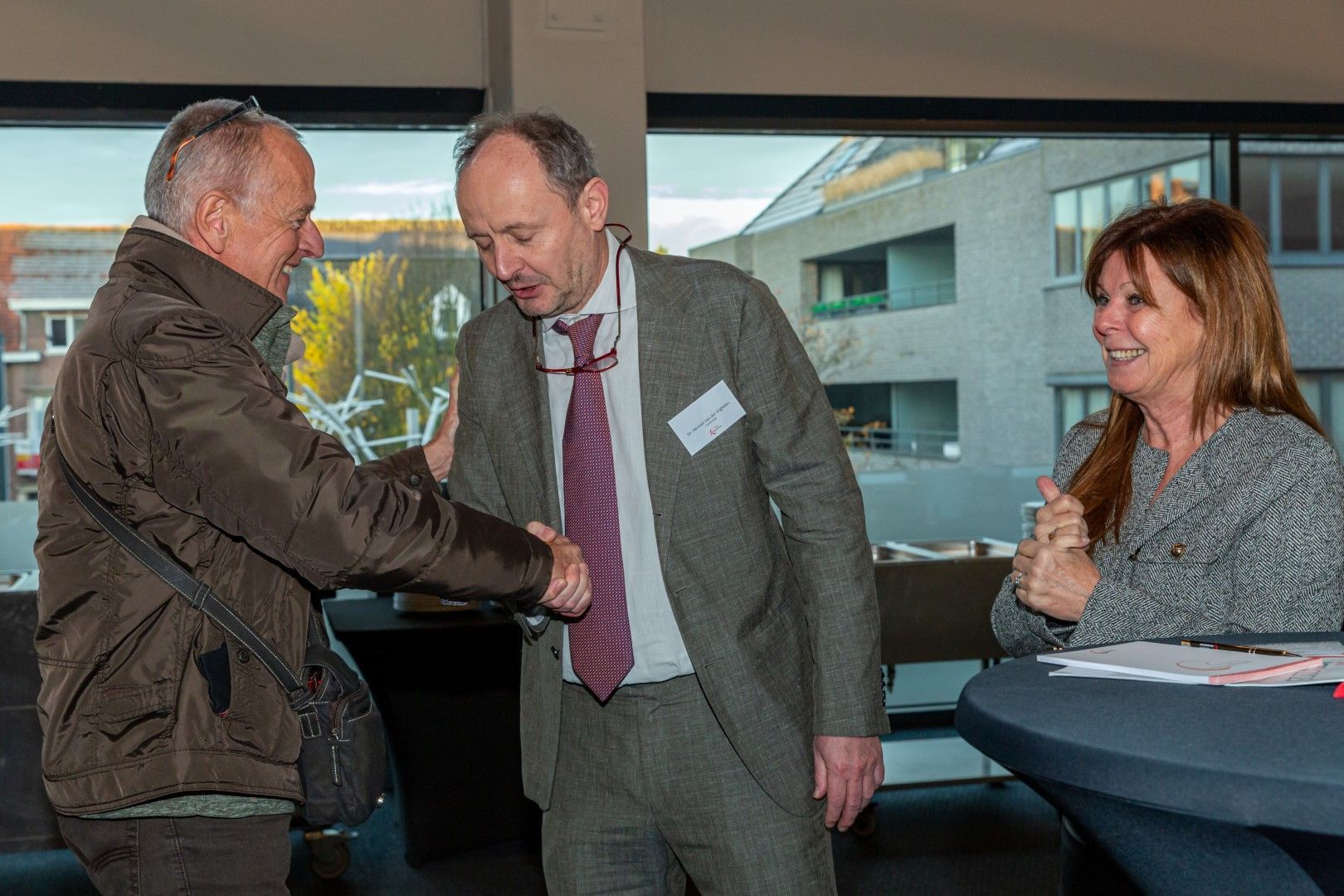 Drie mensen bij een evenement binnenshuis schudden elkaar de hand en glimlachen bij een inschrijftafel.