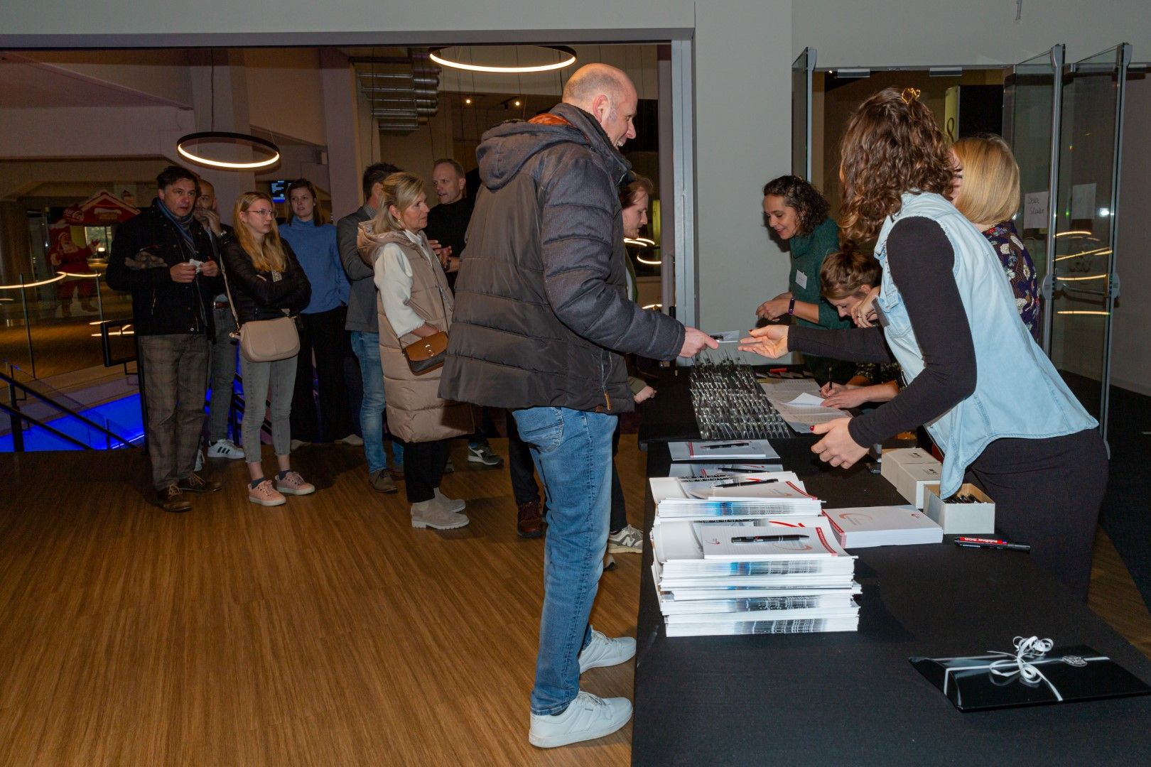 Mensen die in een rij staan bij een tafel met boeken en documenten in een lobby of vergaderzaal.