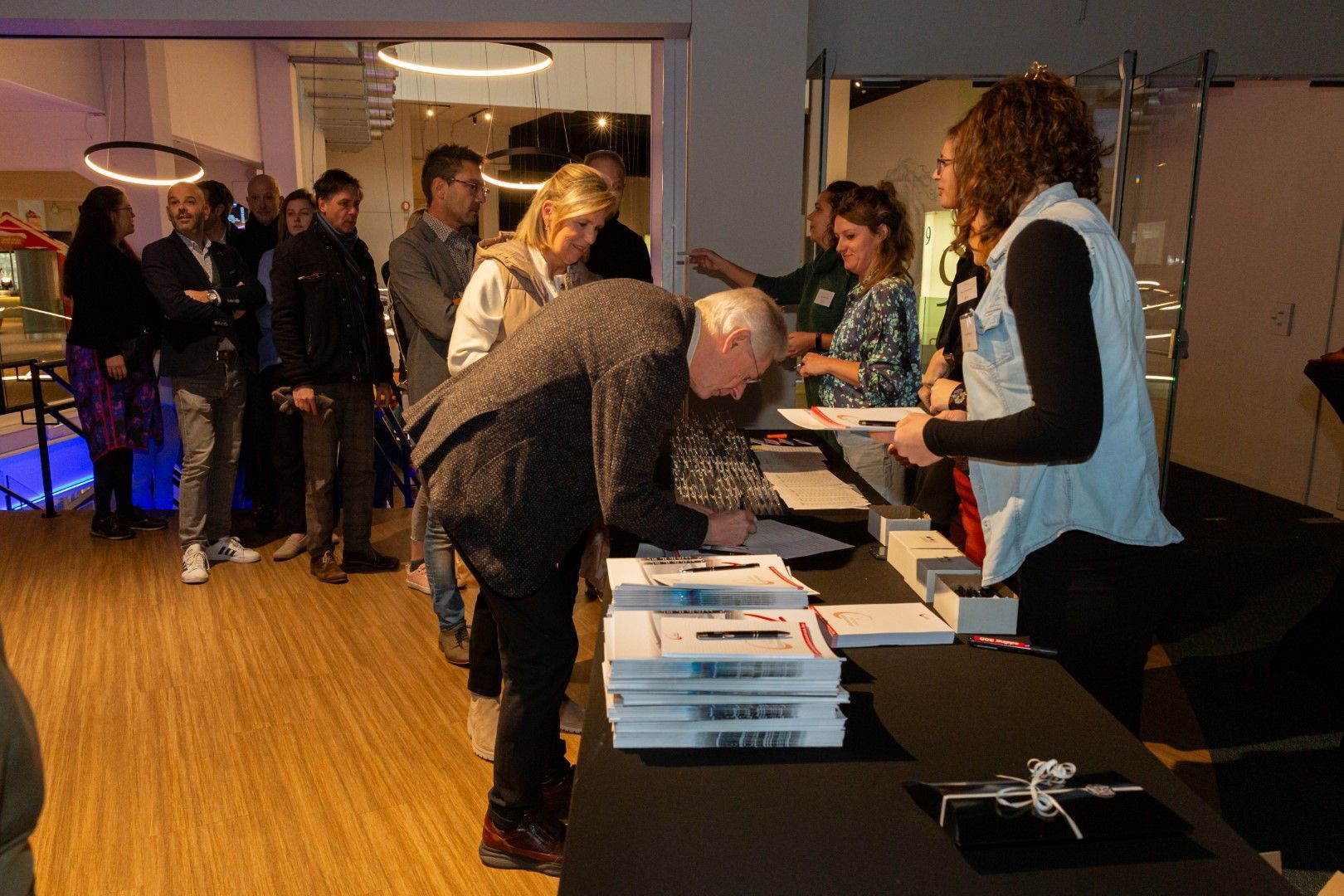 Mensen bij een registratietafel in een lobby, met één persoon die formulieren ondertekent en stapels papier op de tafel.