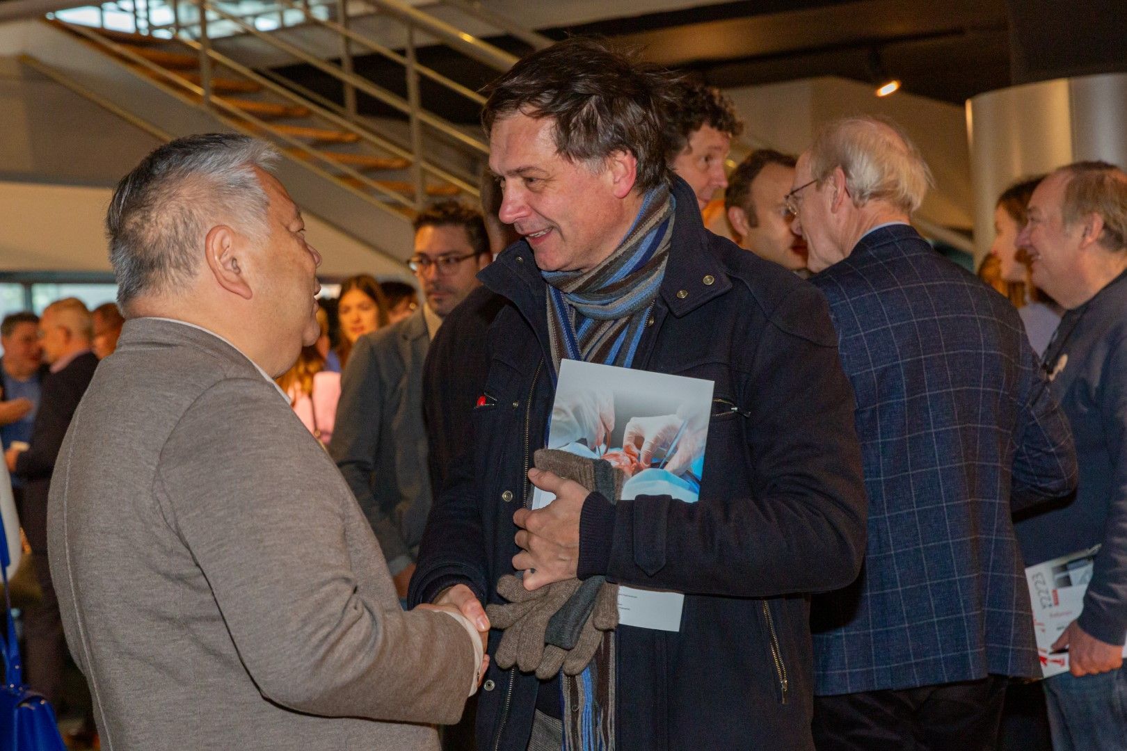 Twee mannen praten met elkaar tijdens een evenement binnenshuis; een van hen houdt een brochure vast, met andere aanwezigen op de achtergrond.