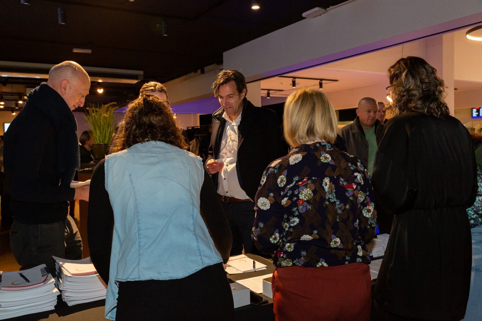 Mensen zaten rond een tafel bij een schemerig verlichte bijeenkomst binnen, te praten en documenten te bekijken.