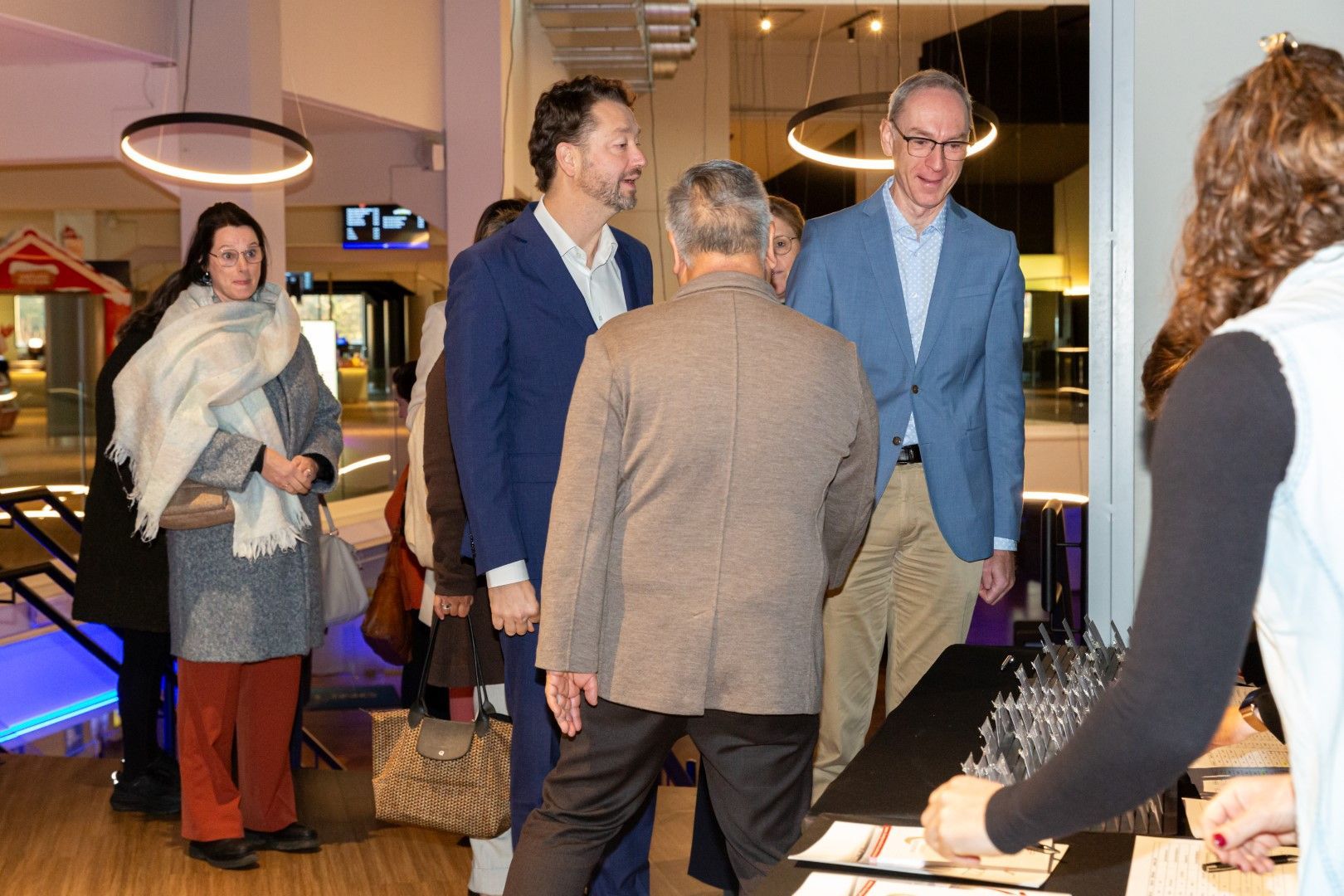 Mensen mengen zich onder elkaar tijdens een sfeervol verlicht evenement binnen, waar drankjes en een incheckbalie zichtbaar zijn.