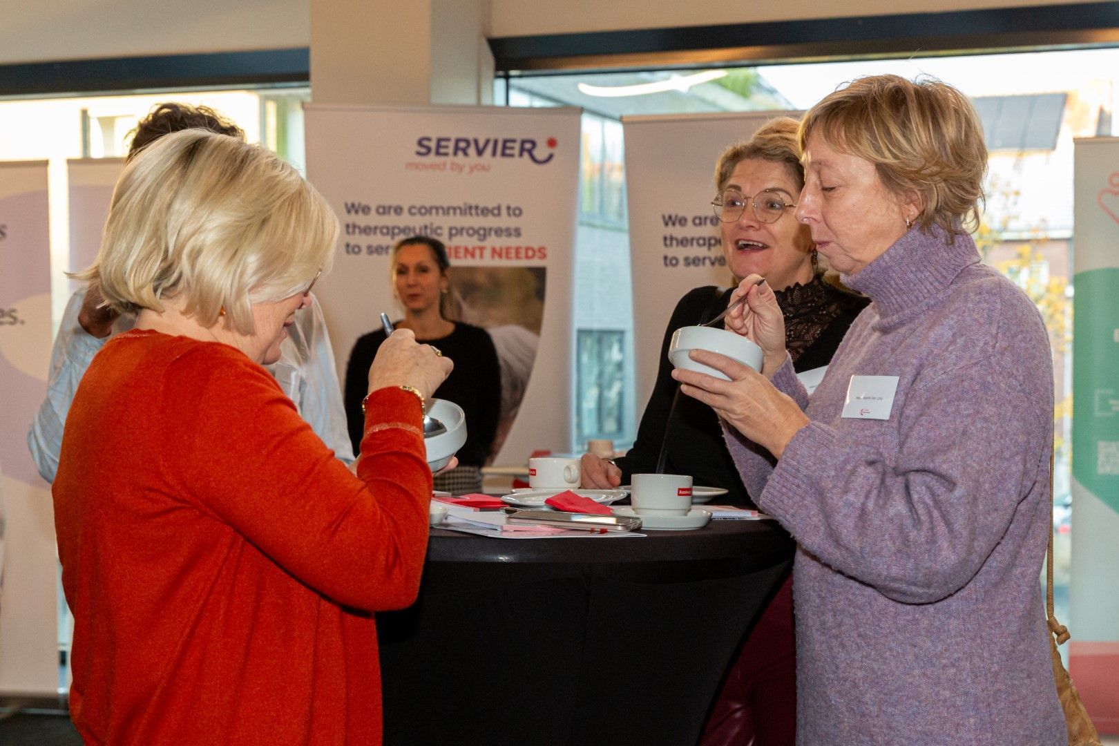 Mensen staan te praten bij een informatiestandje van een buurtvereniging, met koffiekopjes en een bordje met 'SERVIER' op de achtergrond.