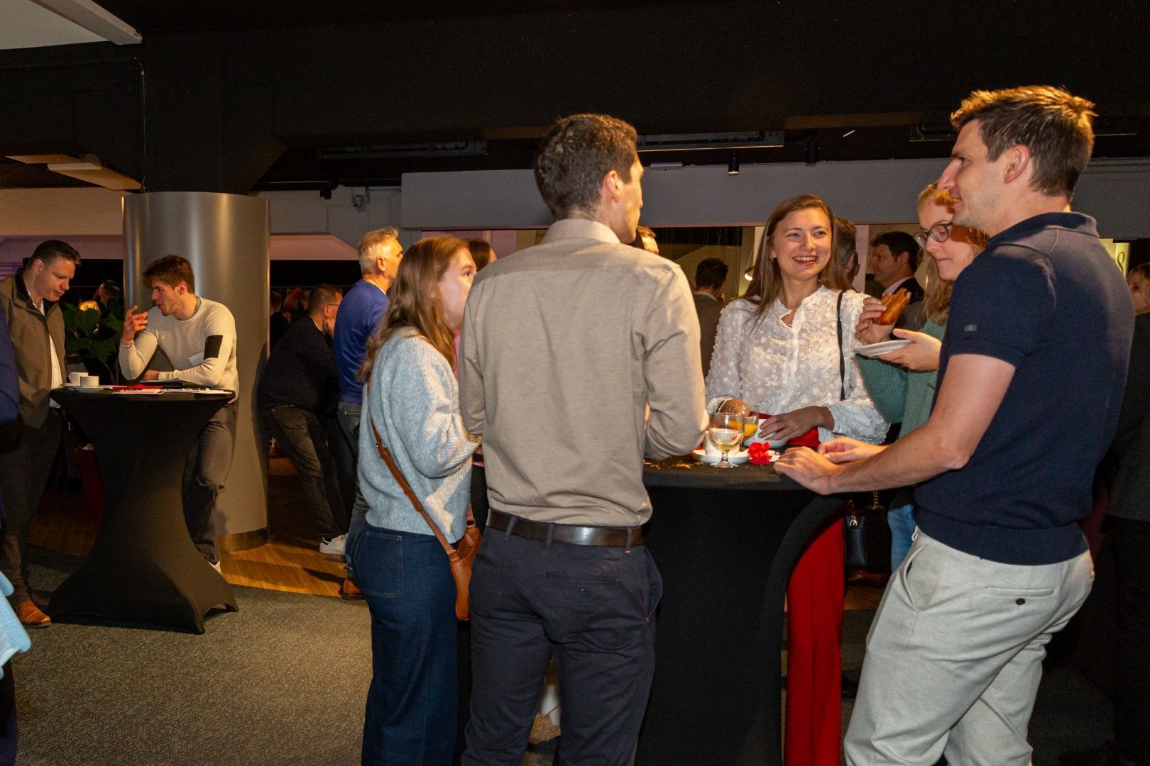 Mensen die met elkaar praten rond een hoge tafel tijdens een schemerig verlichte bijeenkomst binnenshuis.