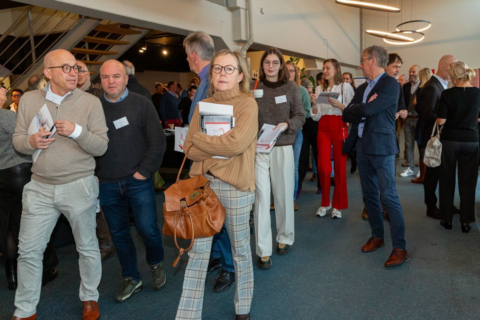 Mensen staan in de rij bij een evenement binnenshuis, te wachten met bekers en tassen.