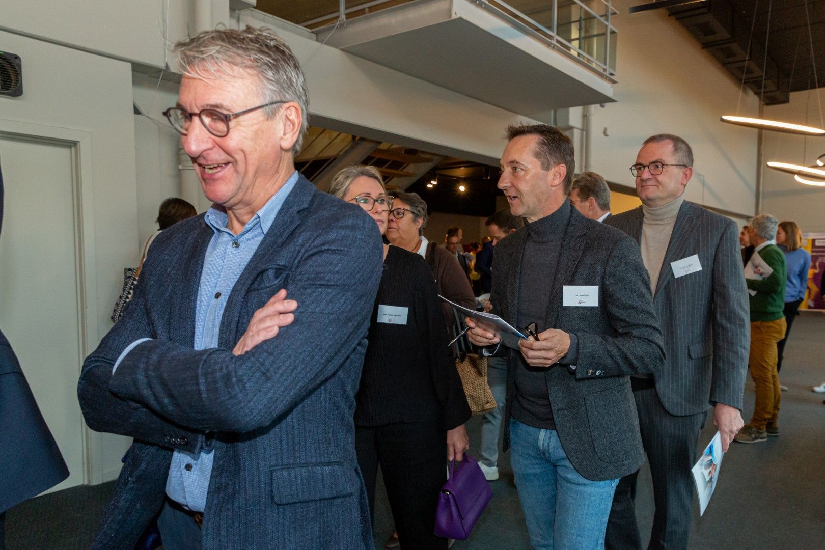 Mensen staan in de rij bij een evenement binnenshuis, met op de voorgrond een lachende man in een blauwe jas.
