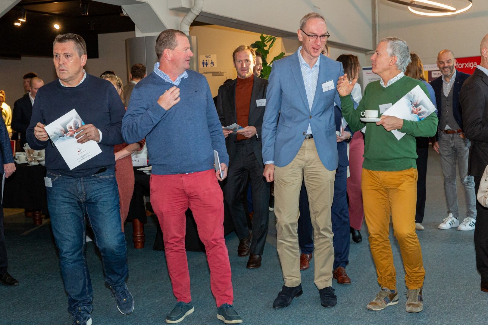 Mensen staan in een lobby, glimlachen en praten terwijl ze papieren en drankjes vasthouden.