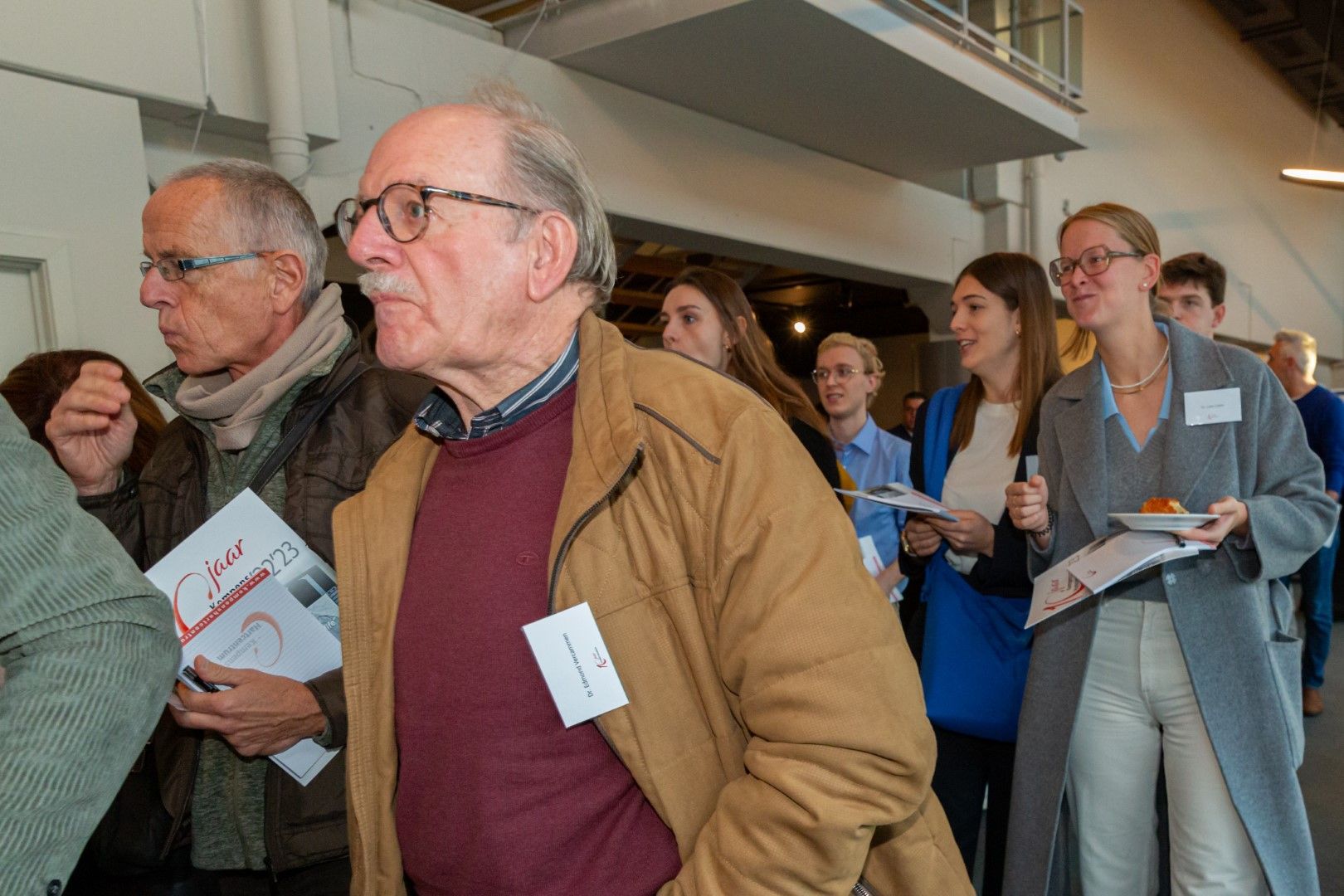 Mensen met flyers lopen door een lichte, overdekte evenementenruimte, aangevoerd door een oudere man in een beige jas.