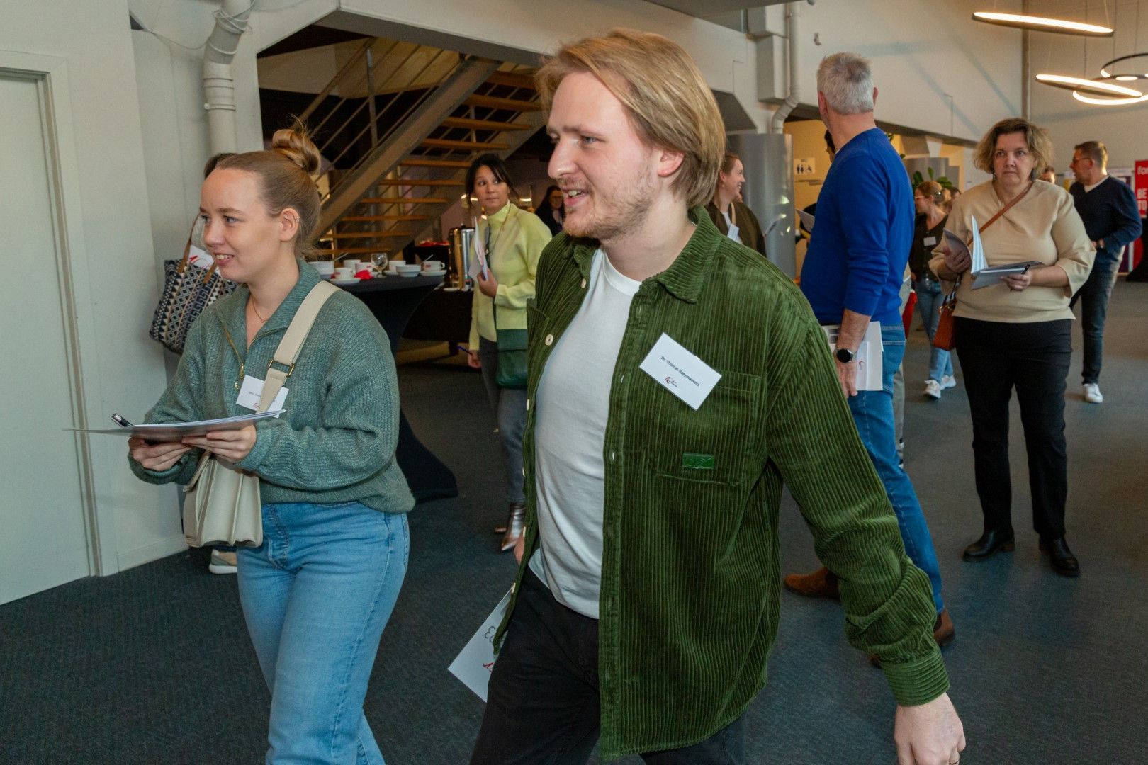 Mensen lopen in een lichte lobby, een van hen met een tablet in de hand en een ander in een groene jas met een glimlach.