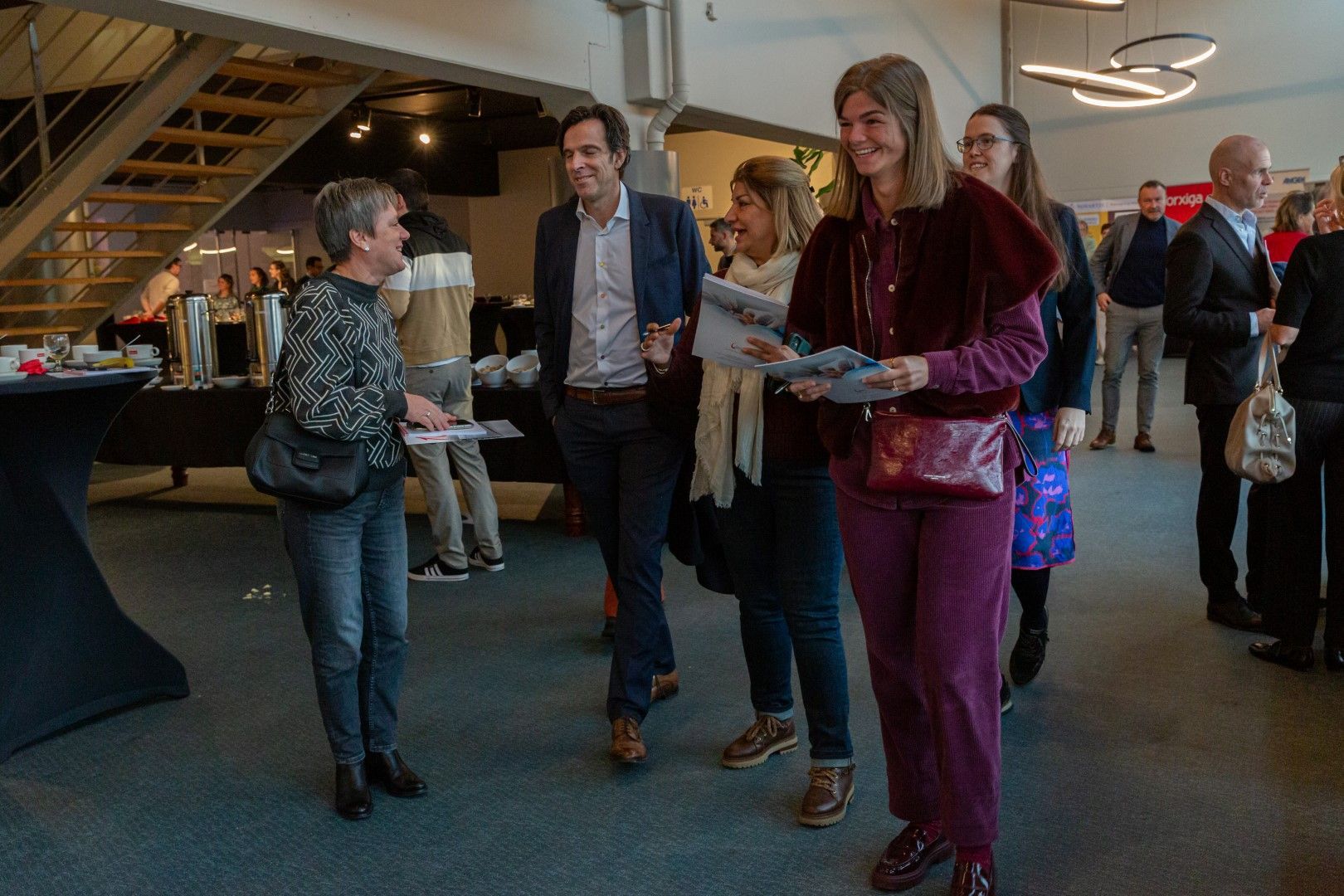 Mensen mengen zich onder elkaar tijdens een evenement binnenshuis, sommigen met drankjes en borden in de hand, onder een trap in een moderne zaal.