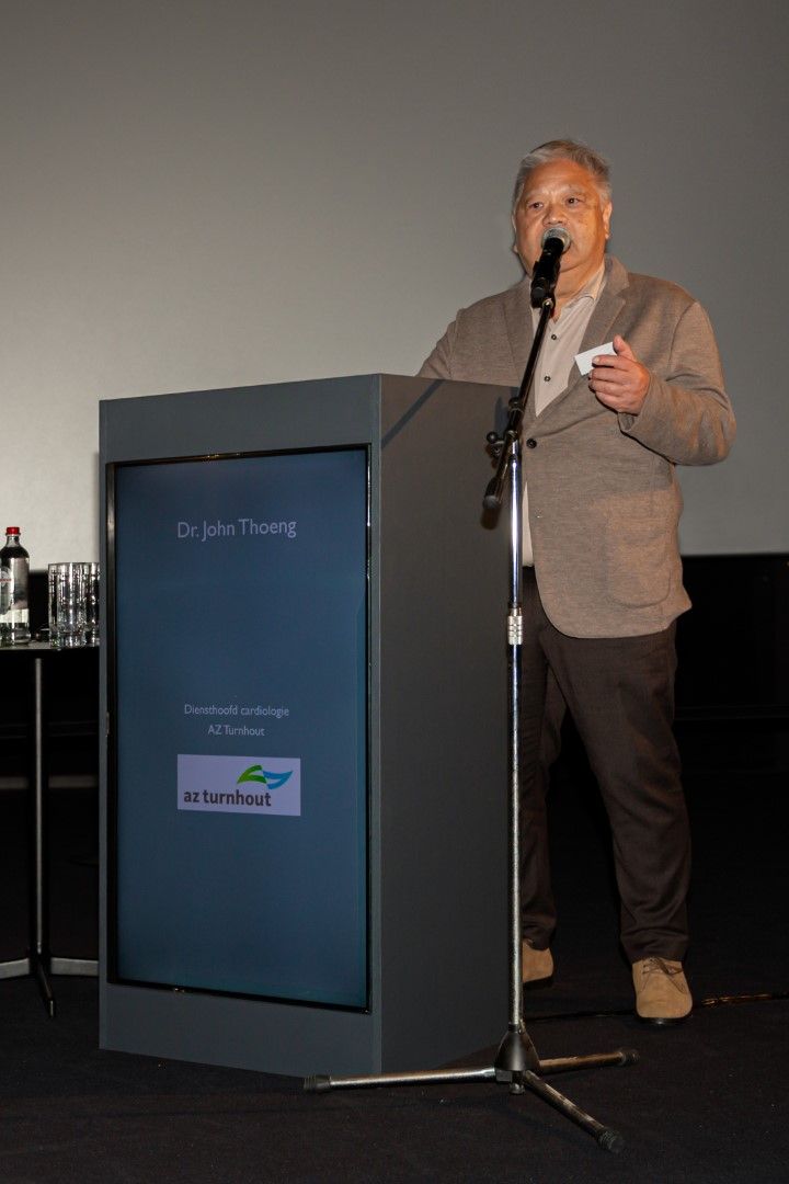 Een man spreekt vanaf een podium op een lessenaar, met een microfoon in zijn hand voor een grijs scherm.