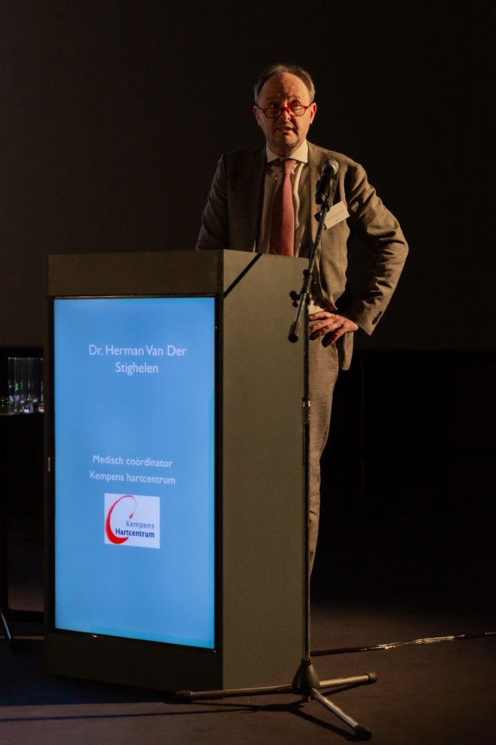 Een man spreekt vanaf een podium met een blauw verlicht frontpaneel op een donker toneel.