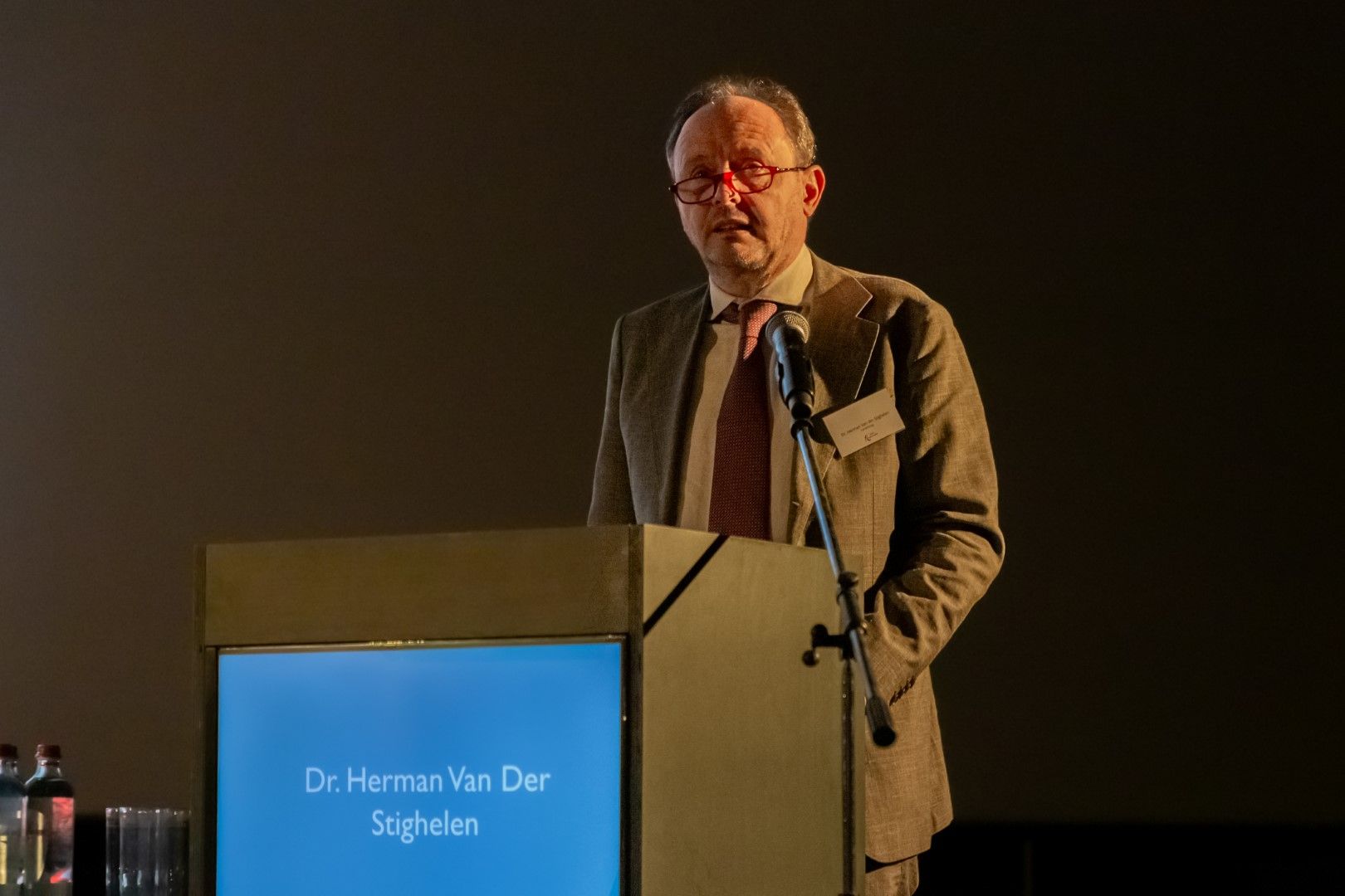Een man spreekt vanaf een podium op een schemerig verlicht toneel, met zijn naam op het voorpaneel.