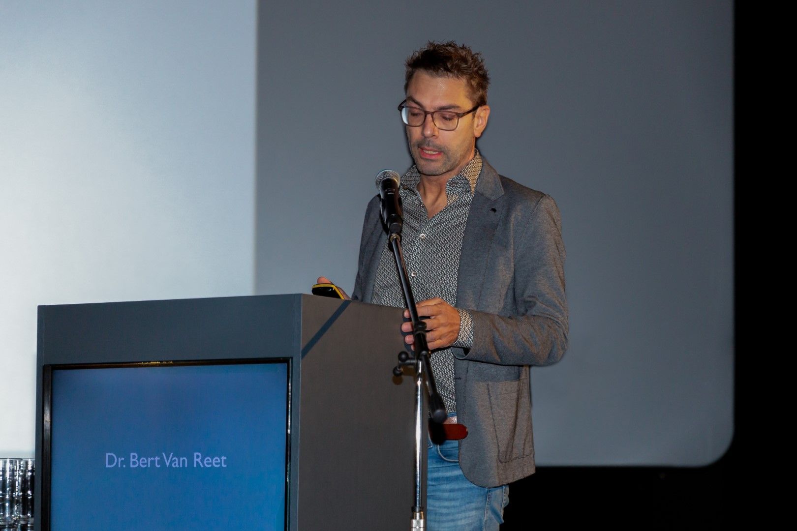 Een man, gekleed in een grijze blazer, spreekt vanaf een podium met een microfoon tijdens een presentatie.