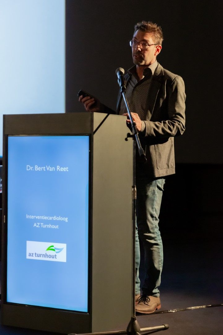 De presentator staat naast een podium en spreekt voor een scherm waarop beelden worden geprojecteerd.