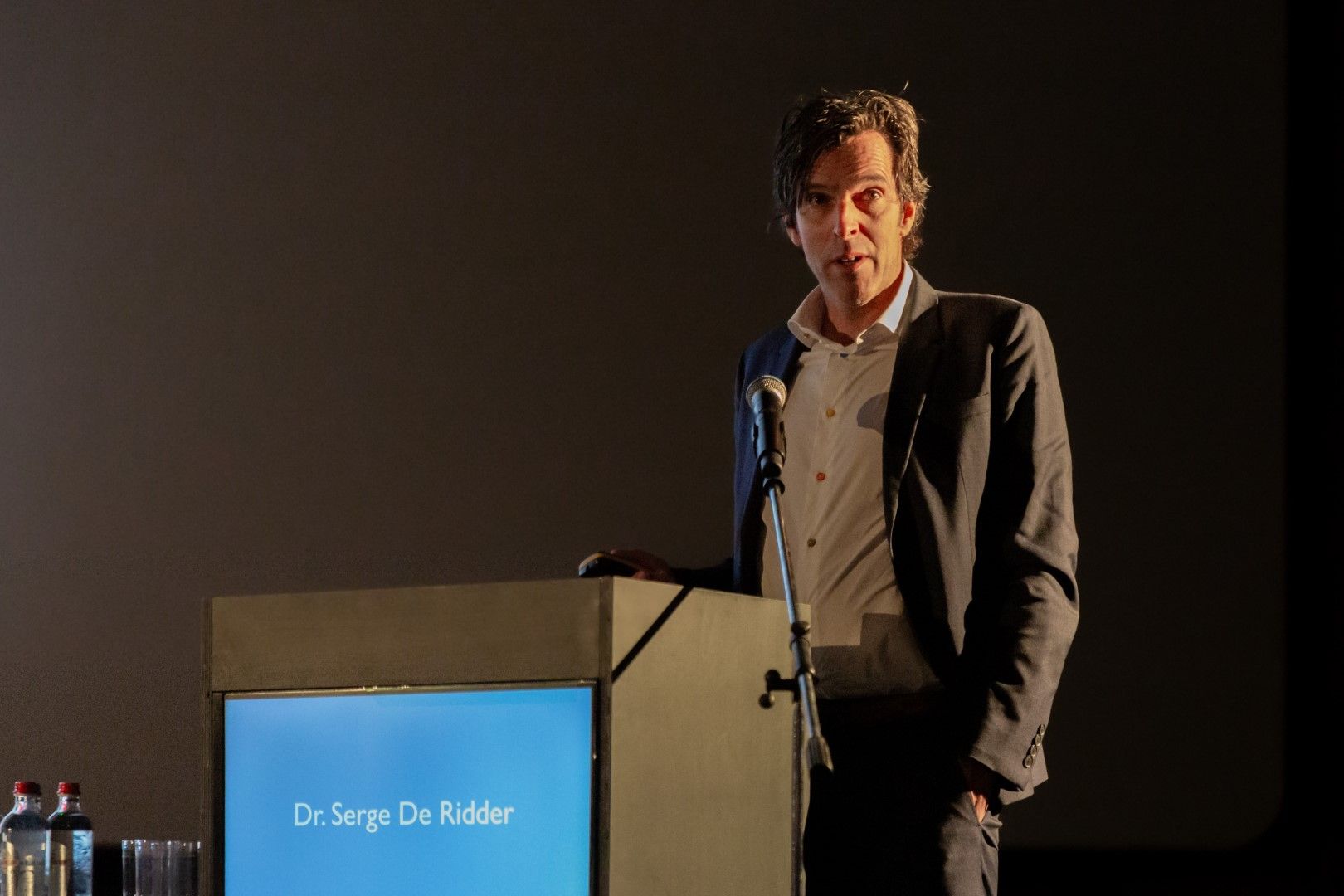 Een man spreekt vanaf een podium op een schemerig verlicht toneel met een blauw naambordje op het scherm.