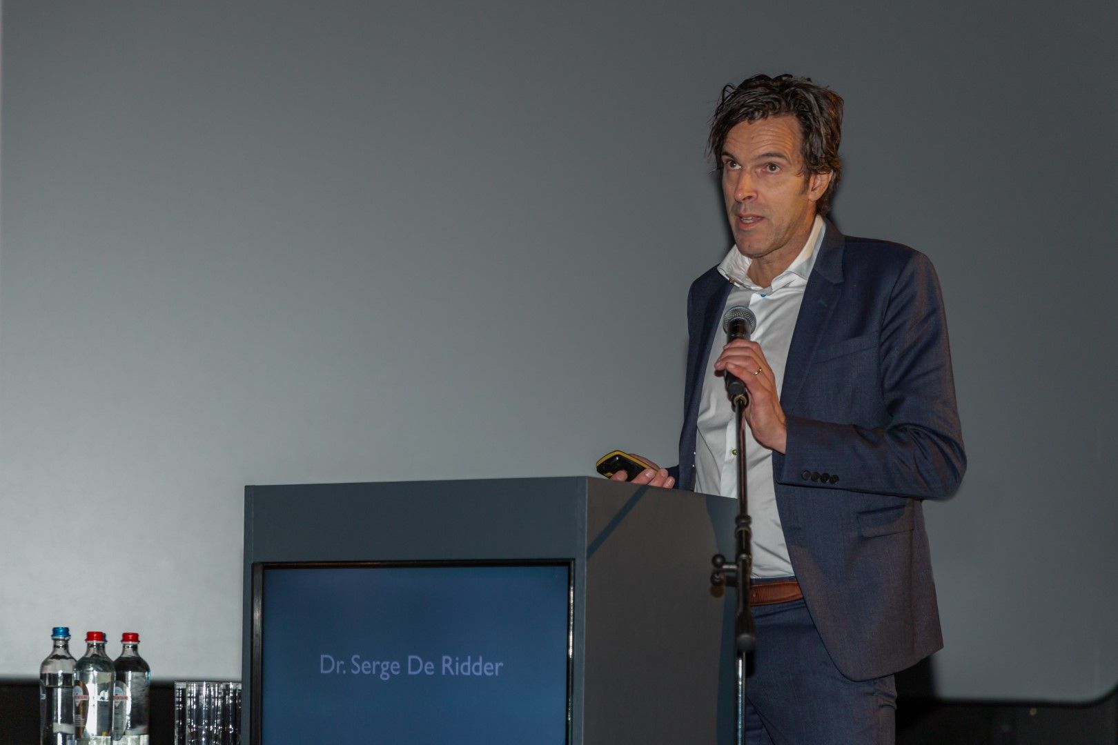 Een man spreekt vanaf een podium met een microfoon tijdens een presentatie in een schemerige vergaderruimte.