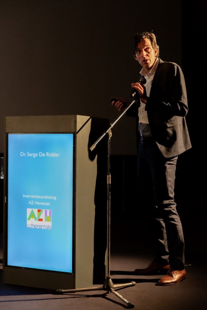 Een man spreekt vanaf een podium met een verlicht blauw scherm, op een donker toneel.