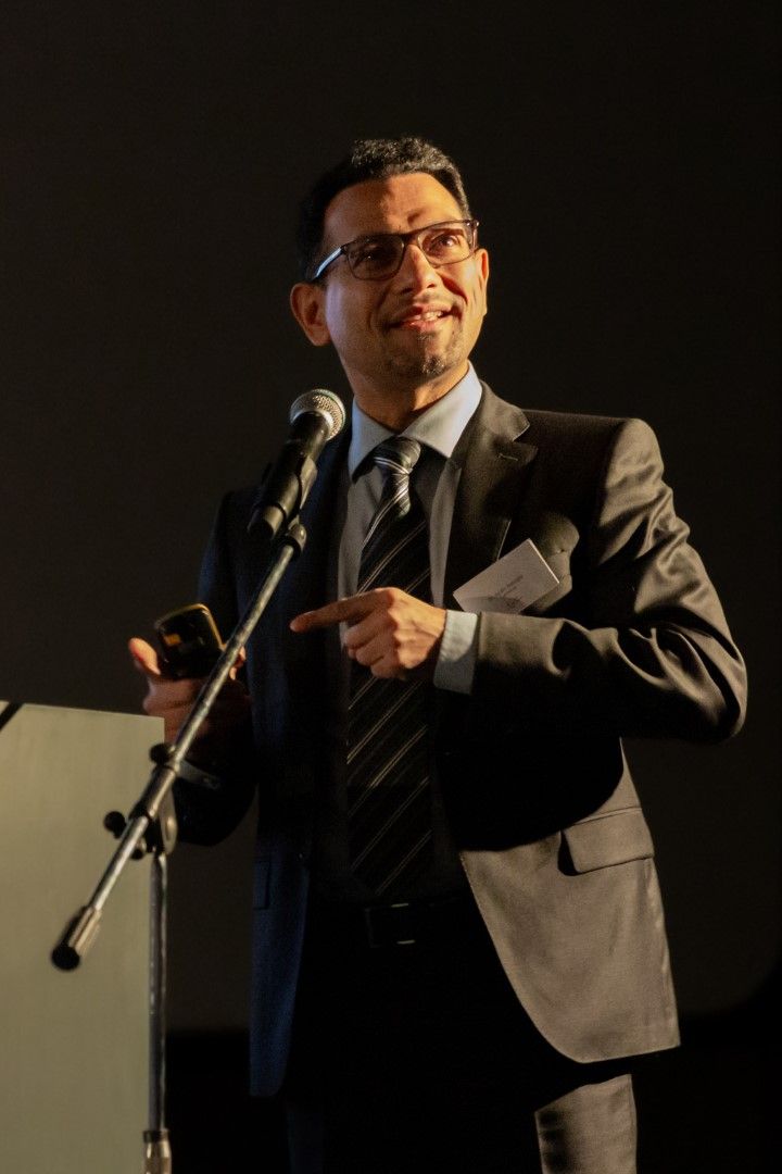 Spreker achter een microfoon op een podium, gekleed in een grijs pak en met een bril op, gebarend tijdens zijn presentatie op het podium.
