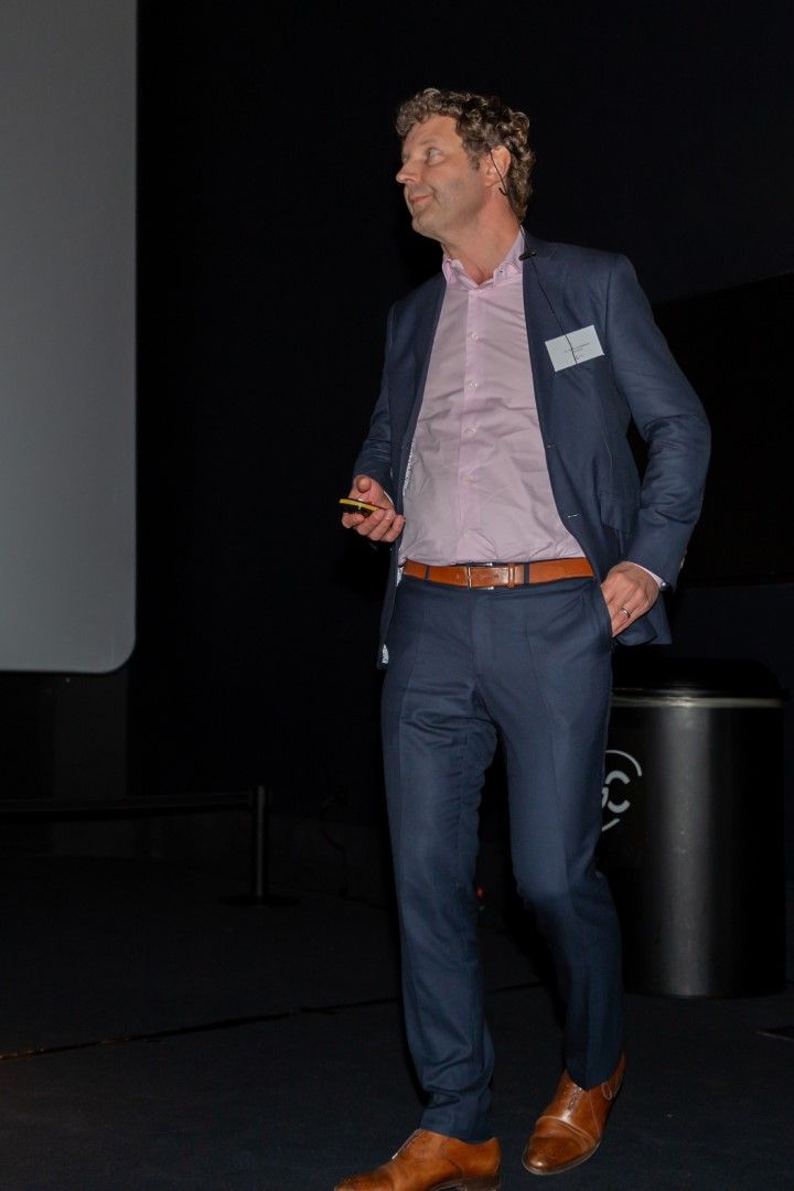 Een man in een blauw pak staat op het podium te spreken, houdt een clicker vast en kijkt opzij.