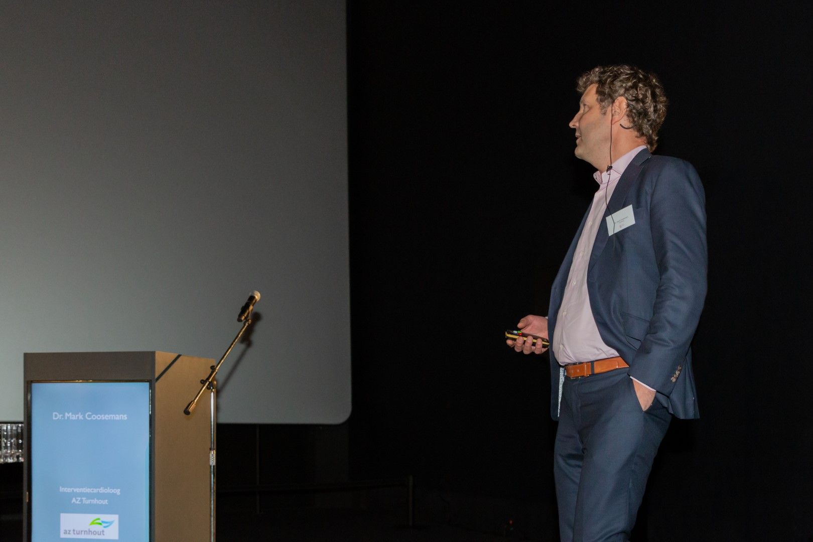 Een man in een blauwe blazer spreekt op een donker podium naast een lessenaar en microfoon.