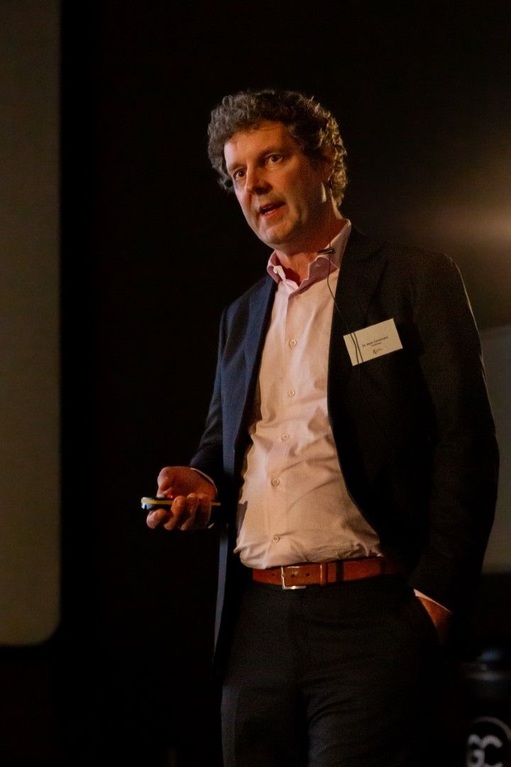 Een man in een donkere blazer en een roze shirt spreekt op een podium, met een microfoon in zijn hand onder warm licht.
