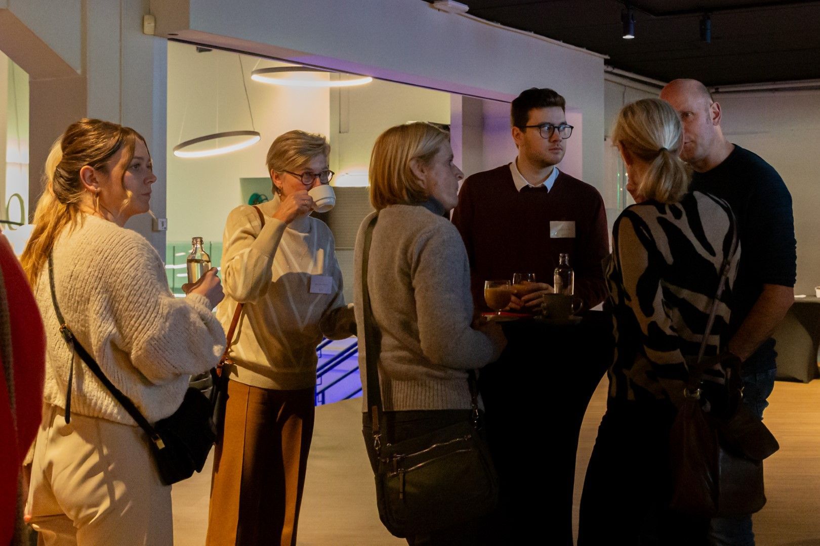 Mensen die in een schemerig verlichte binnenruimte met elkaar praten en drankjes in de hand bij een deuropening vasthouden.