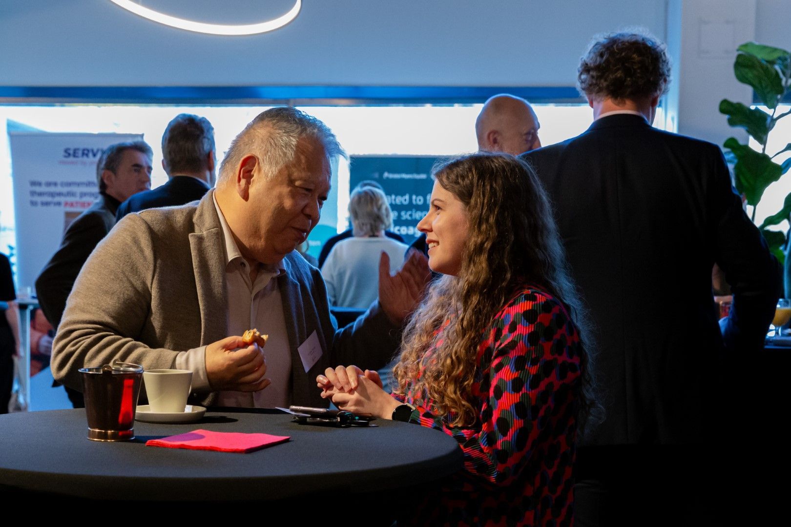 Twee mensen praten aan een ronde tafel tijdens een netwerkevenement, met anderen op de achtergrond.