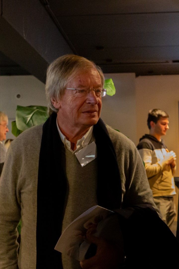 Een man in een donkere trui en een wit overhemd houdt papieren vast in een schemerig verlichte bijeenkomst binnen.