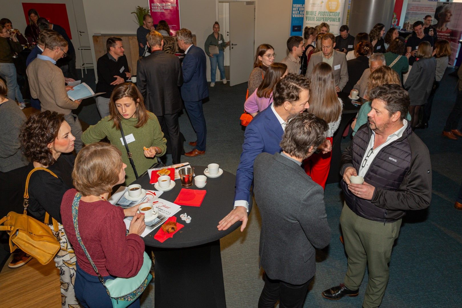 Mensen die tijdens een netwerkevenement binnenshuis met elkaar in gesprek zijn rond tafels met hapjes en drankjes.