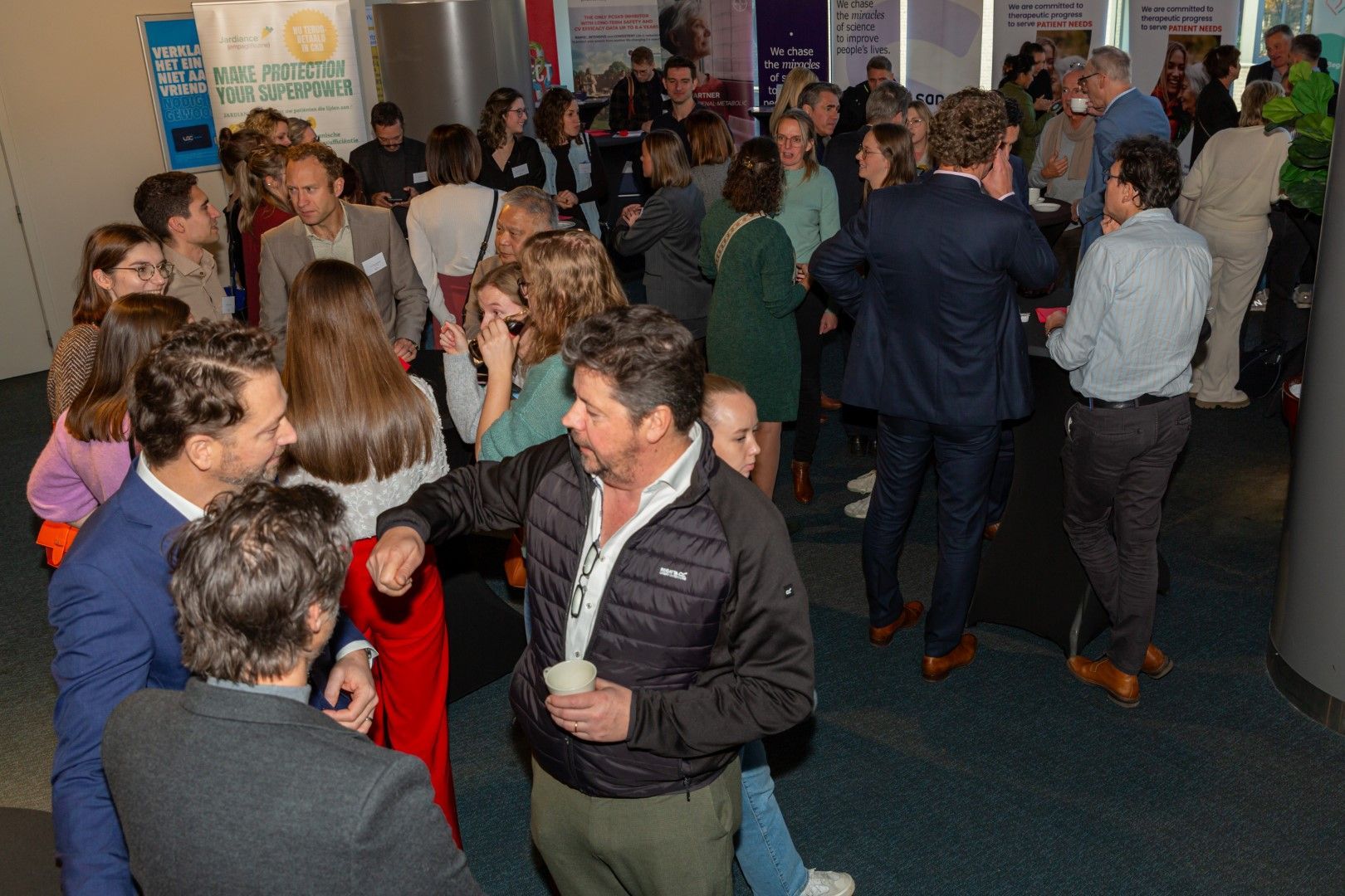 Een drukke ontvangstzaal binnen, waar mensen zich tussen de tafels en banners begeven en met elkaar praten.