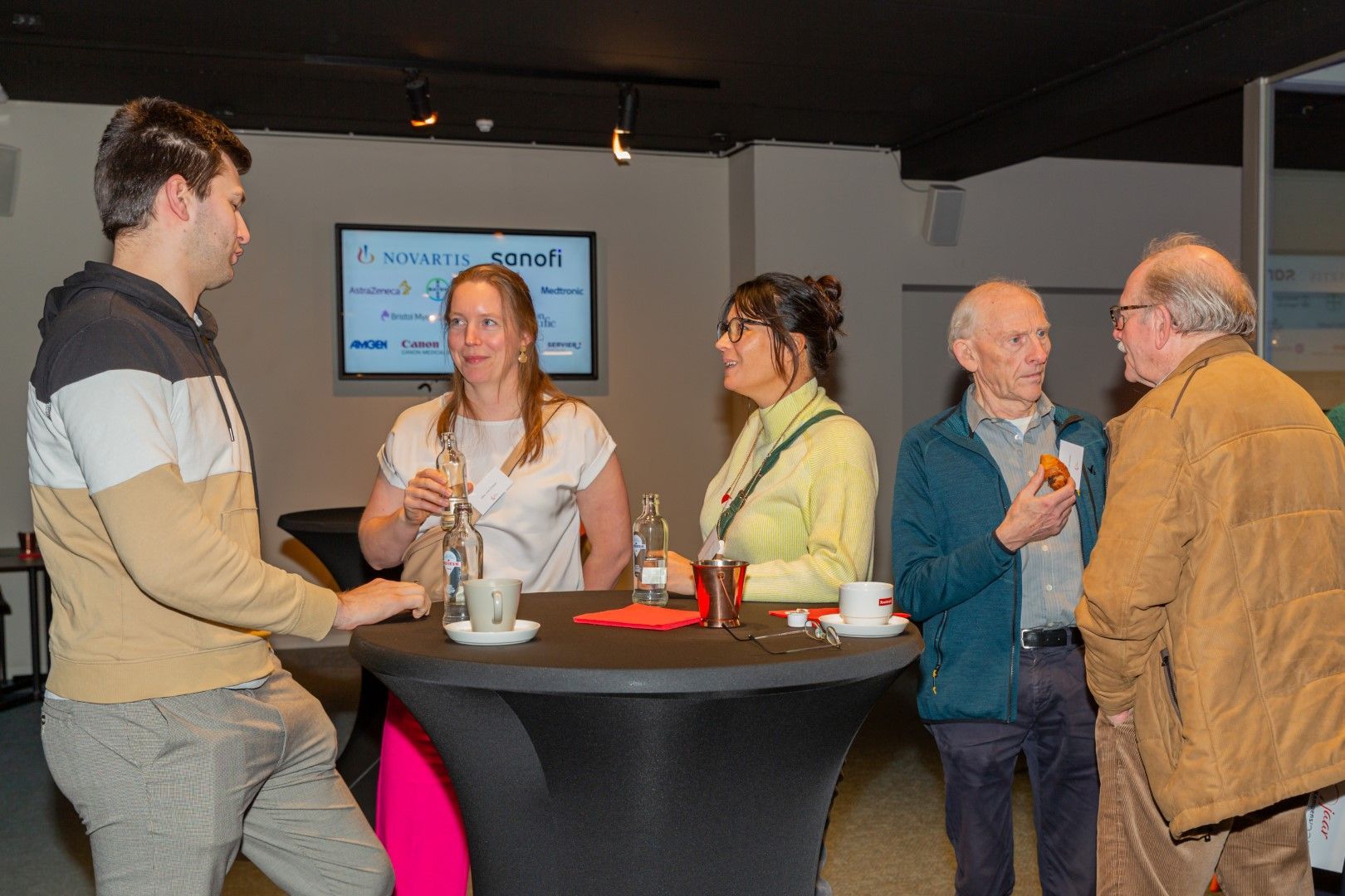 Mensen praten met elkaar rond een hoge tafel in een schemerige lounge, met drankjes en een presentatiescherm op de achtergrond.