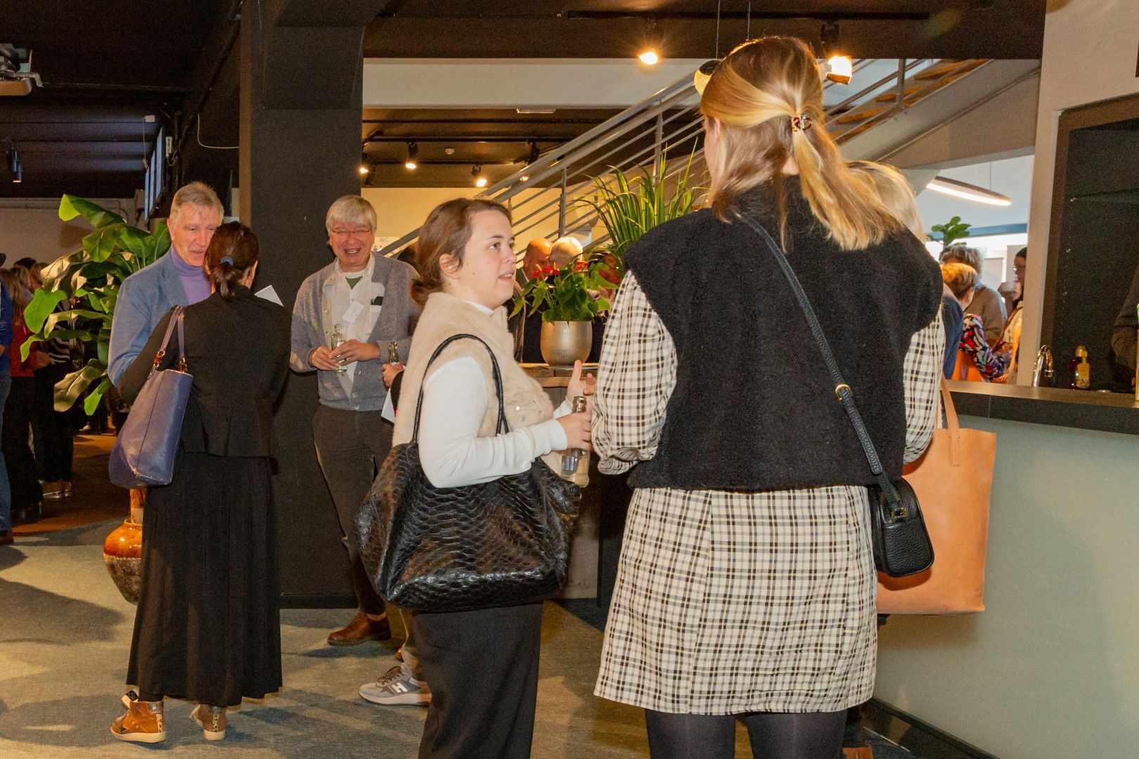 Mensen praten met elkaar in een moderne, overdekte lobby met een houten balie en warme verlichting.