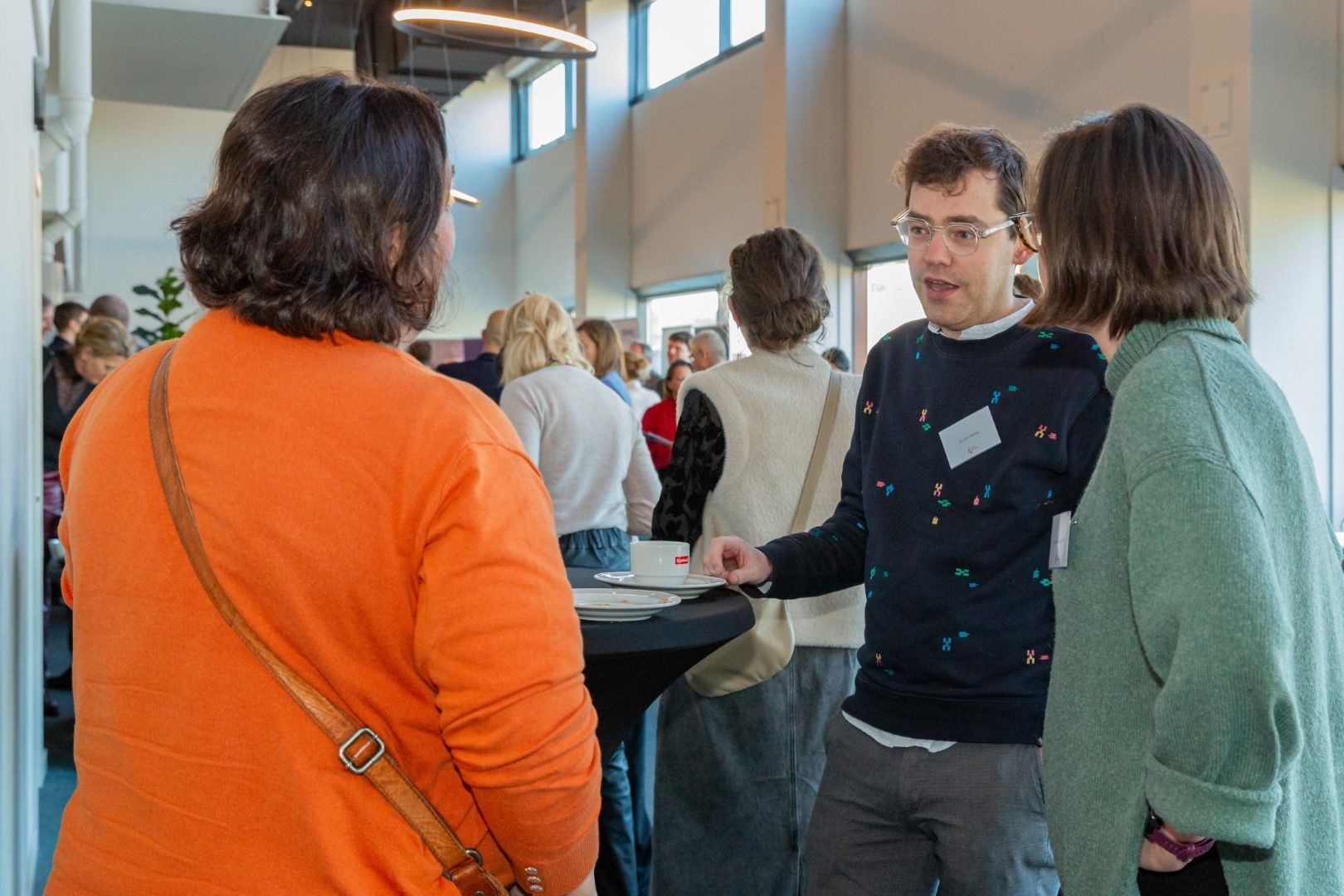 Mensen praten met elkaar tijdens een netwerkevenement binnen, waarbij een man in gesprek is met twee anderen bij hoge ramen.