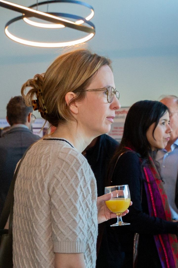Vrouw met bril die een geel drankje vasthoudt tijdens een drukbezocht evenement binnenshuis onder moderne ringverlichting.