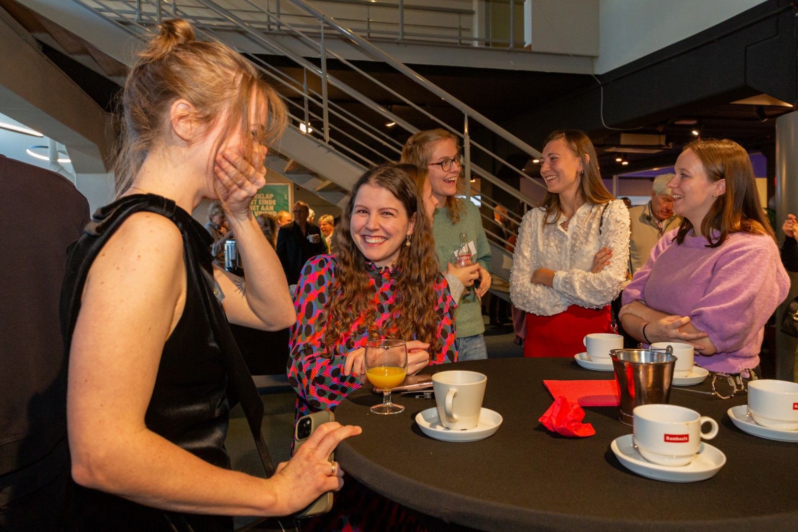Vijf mensen zitten gezellig te praten rond een cafétafel met koffiekopjes en rode servetten in een lichte, overdekte lounge.