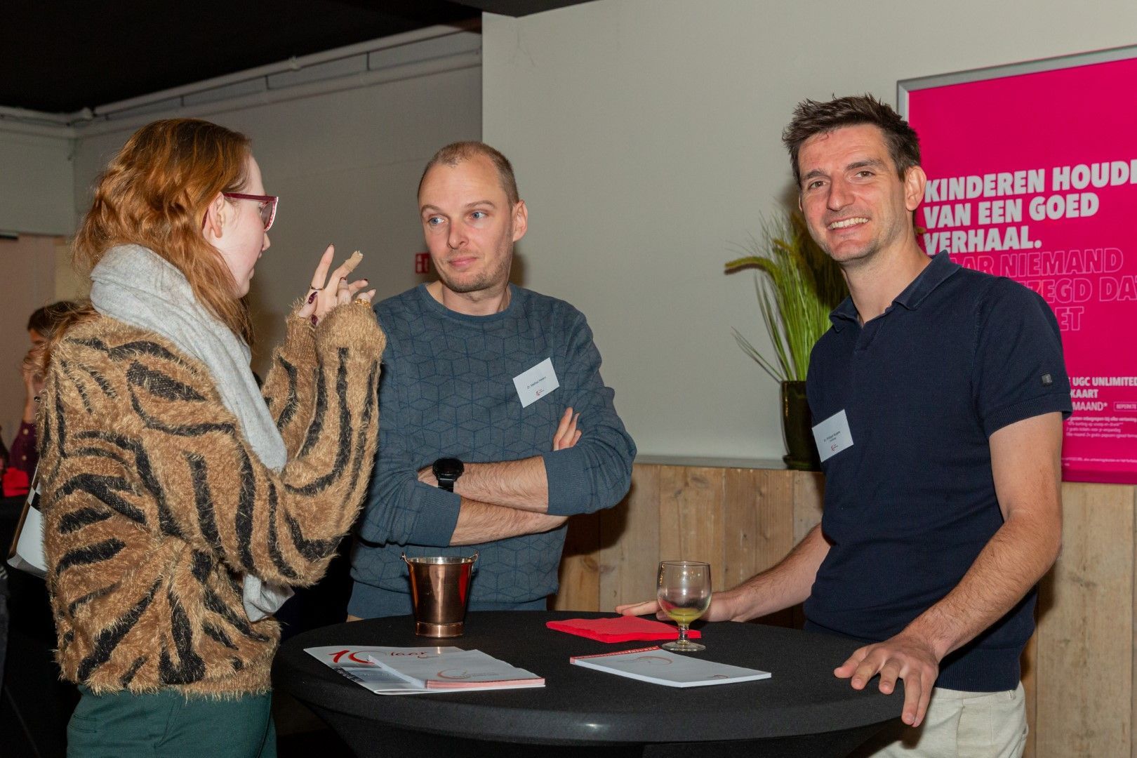 Drie mensen praten rond een kleine ronde tafel tijdens een evenement binnenshuis, met drankjes en kranten.