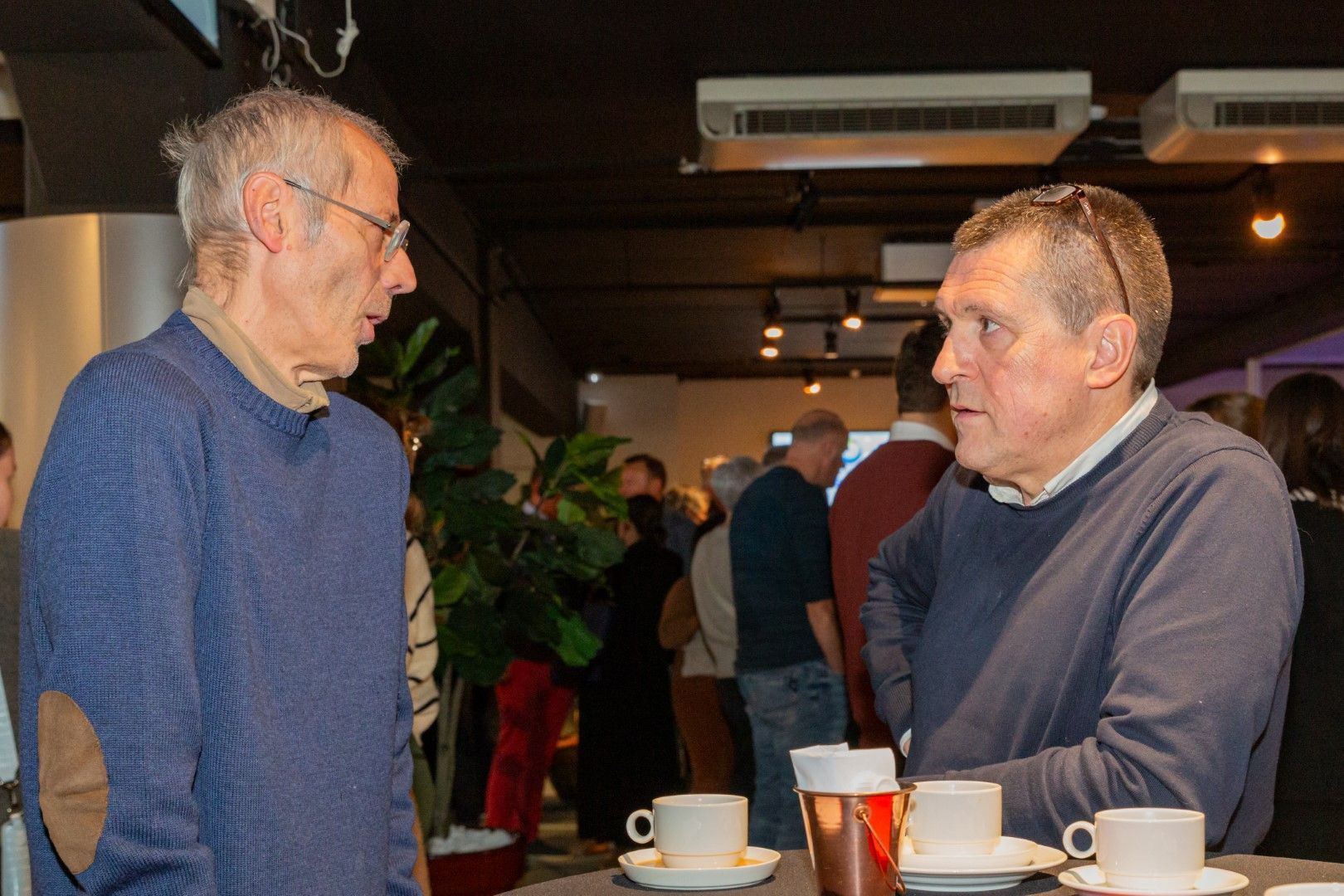Twee mannen praten aan een cafétafel met koffie en een biertje in een schemerig verlichte ruimte.