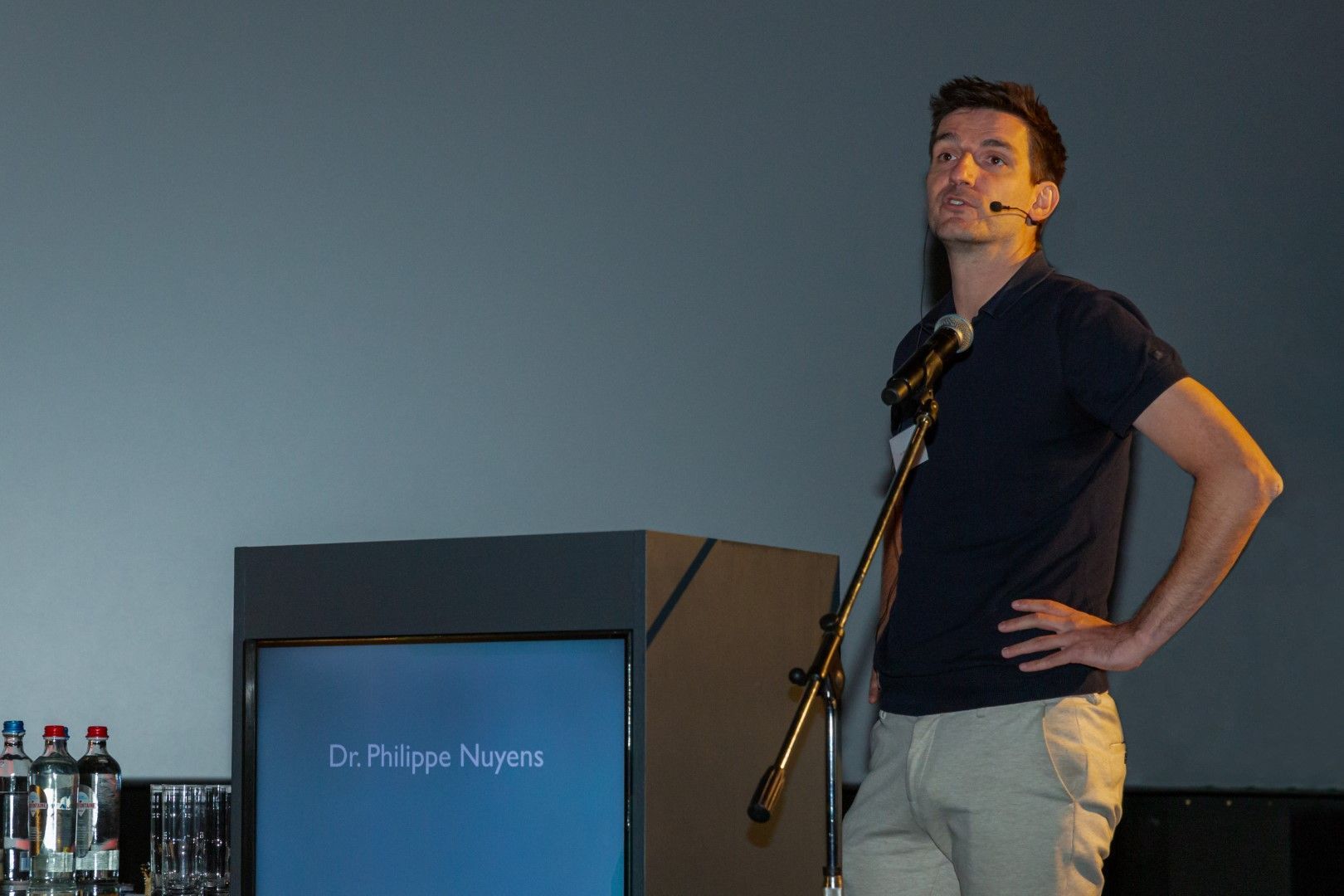 Een man staat achter een podium met een microfoon, naast een scherm waarop